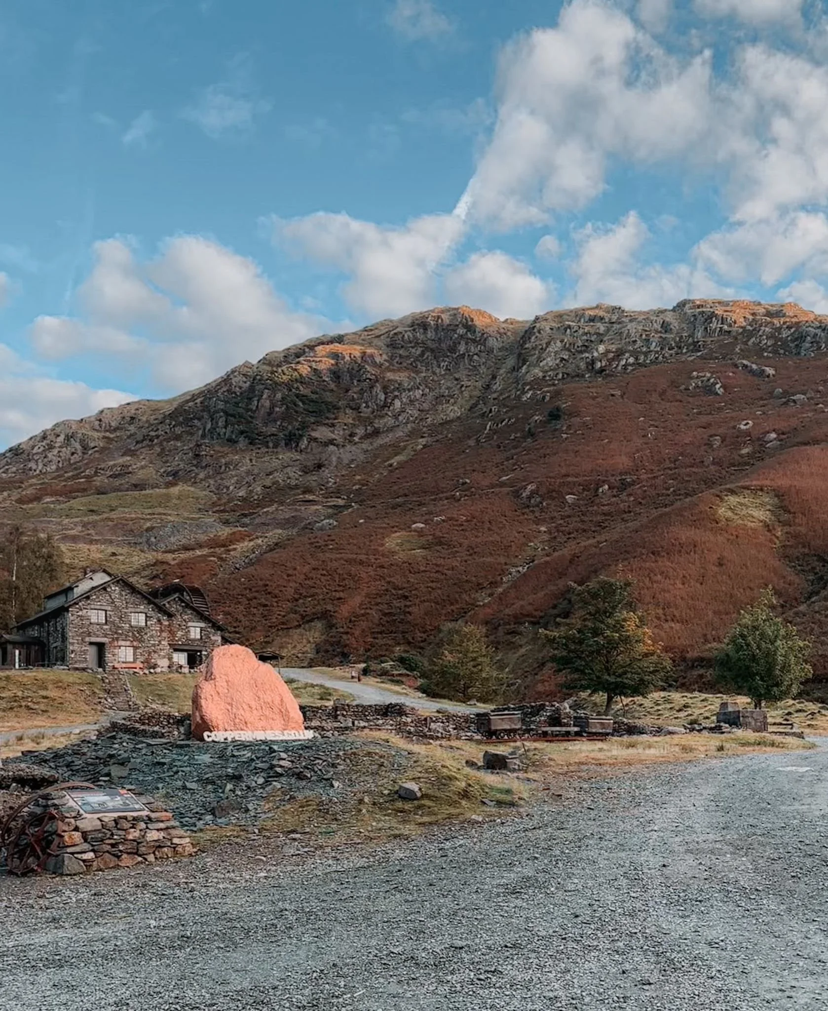 A landscape scene featuring a mountain with rocky slopes and sparse vegetation under a partly cloudy sky. In the foreground, there is a gravel area with a small stone house, a large pink rock, and some scattered stones and debris, including old car parts.