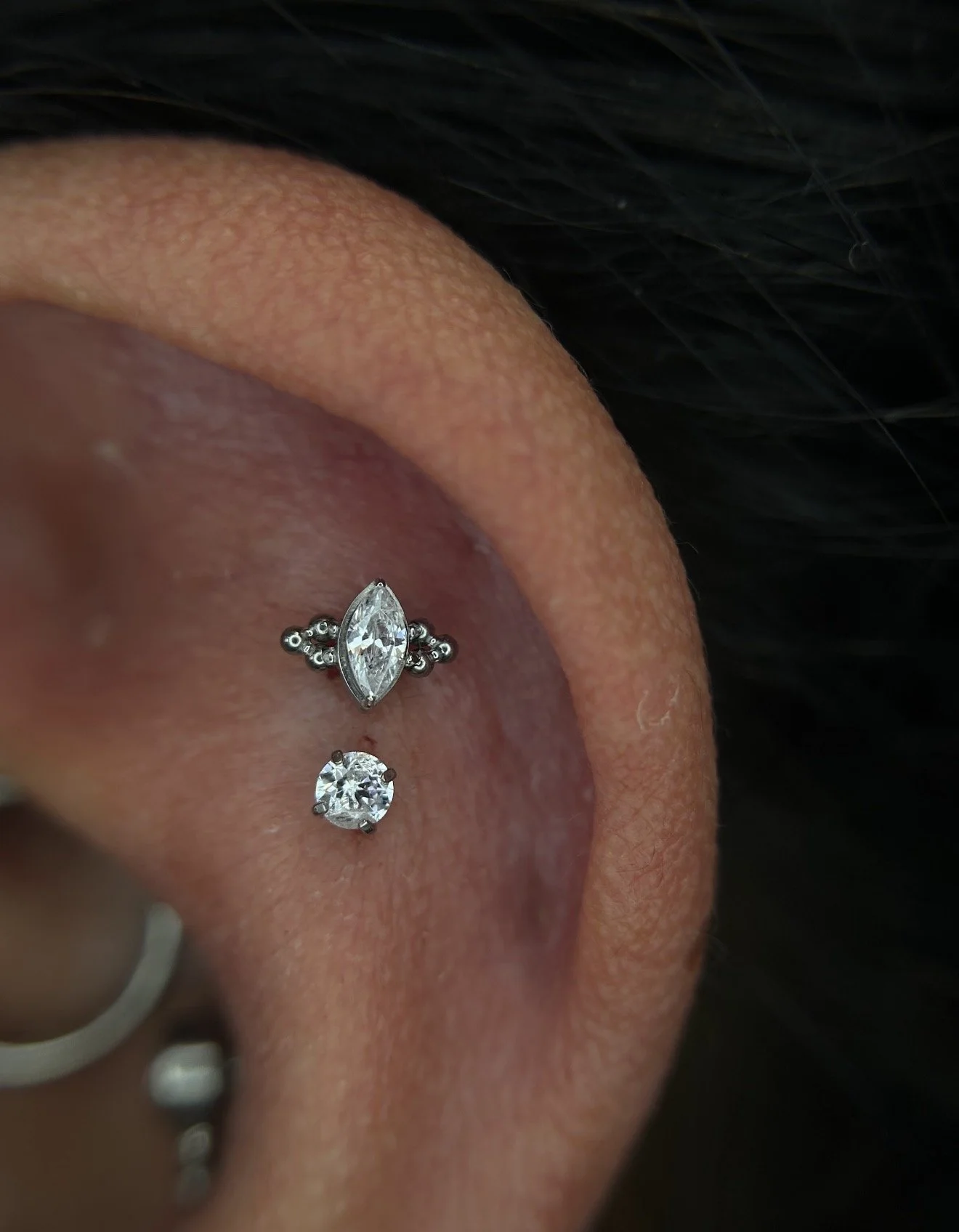 Close-up of a person's ear with a double cartilage piercing, featuring dainty earrings with small clear gemstones, on a background of dark hair.