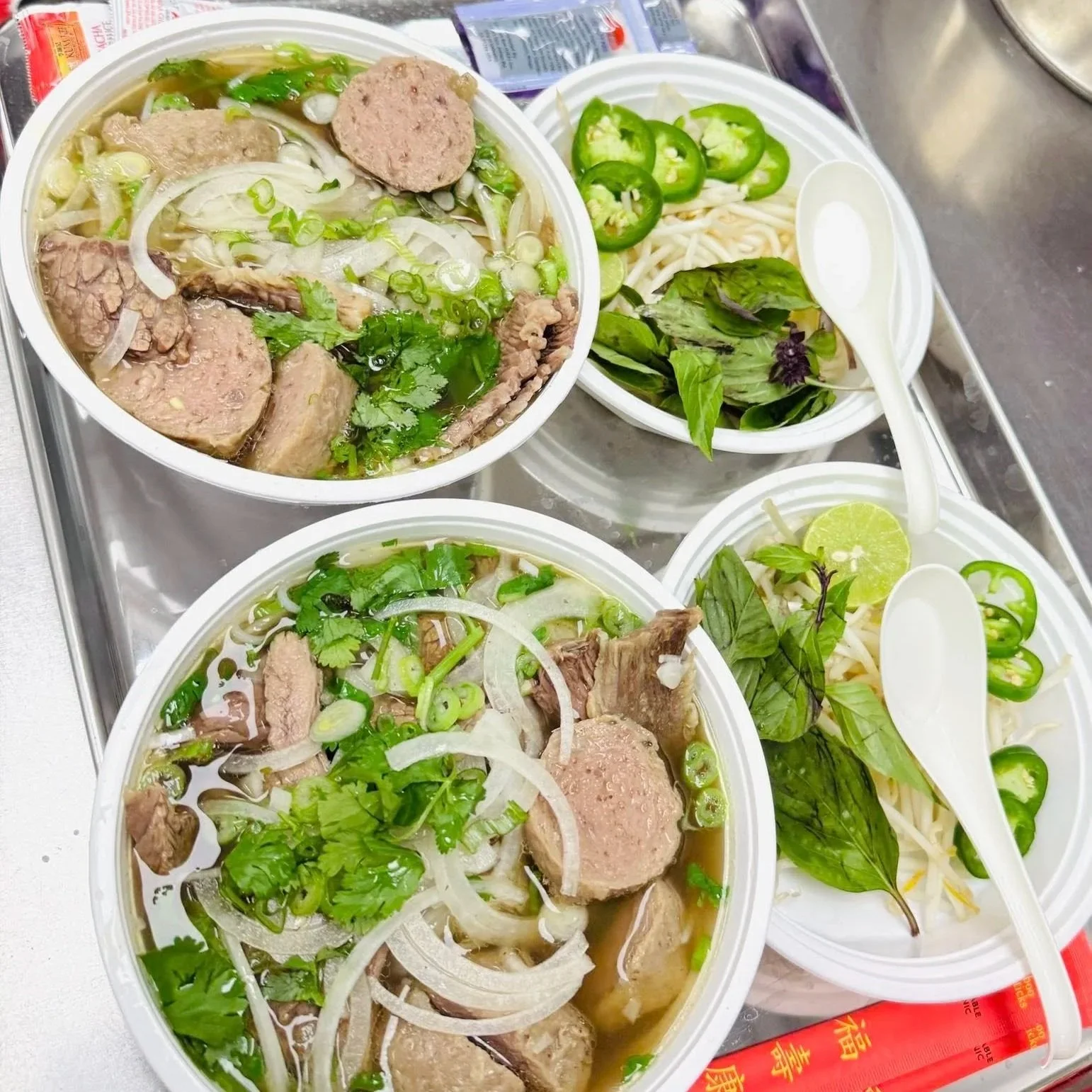 Two bowls of Vietnamese pho with beef meatballs, sliced onions, green herbs, and chopped green onions, along with a side bowl of fresh bean sprouts, sliced green chilies, lime wedges, and herbs, all on a metal tray.