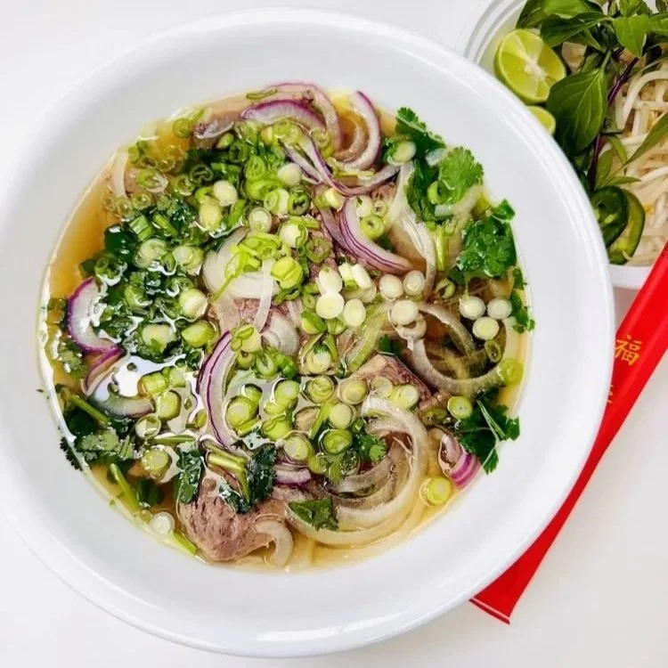 A bowl of Vietnamese pho with sliced beef, chopped green onions, cilantro, and sliced onions in broth, with lime, herbs, and chopsticks on the side.