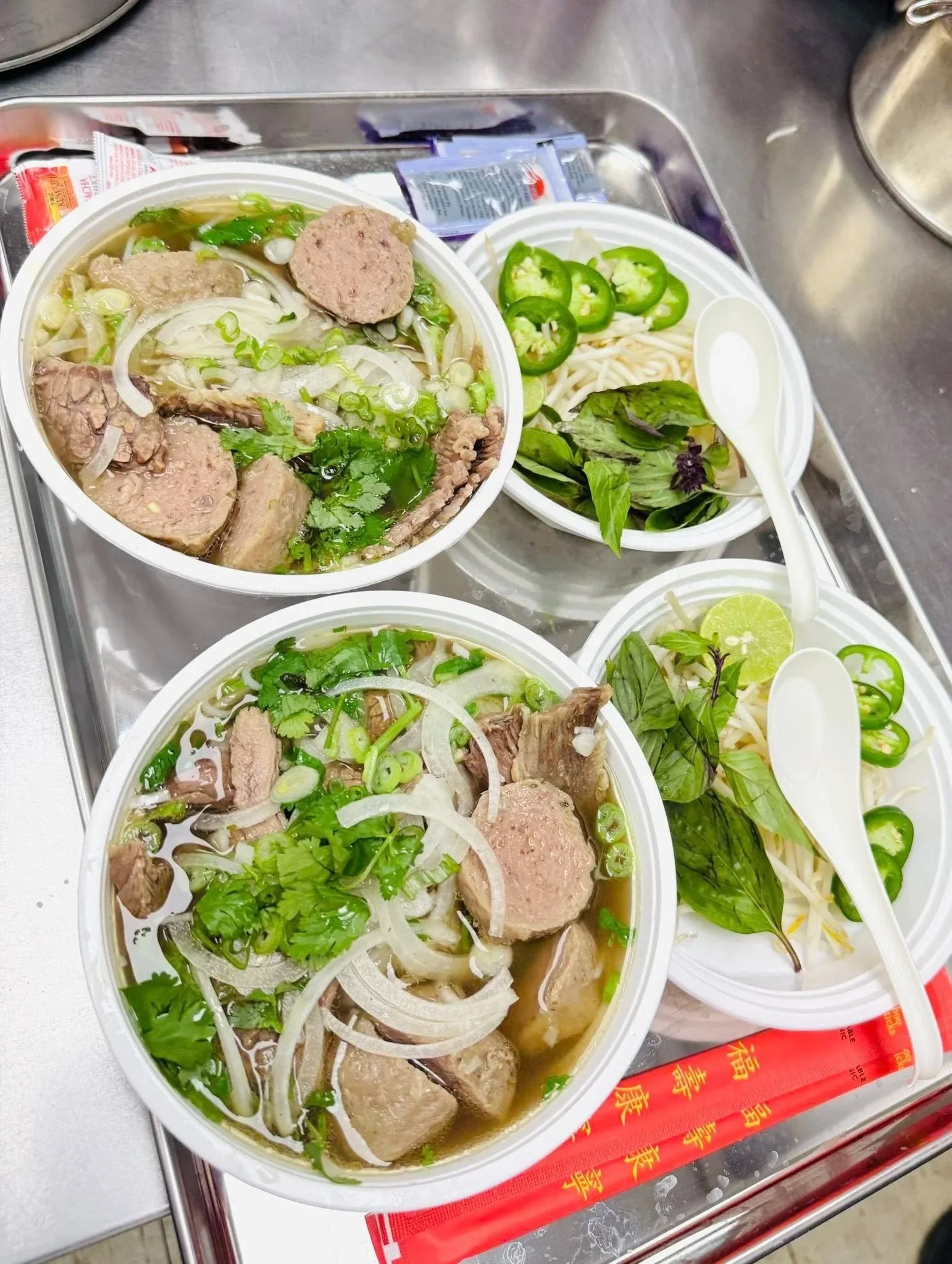 Three bowls of Vietnamese pho with meatballs, noodles, fresh herbs, and lime slices, on a metal tray with chopsticks and packets of condiments.