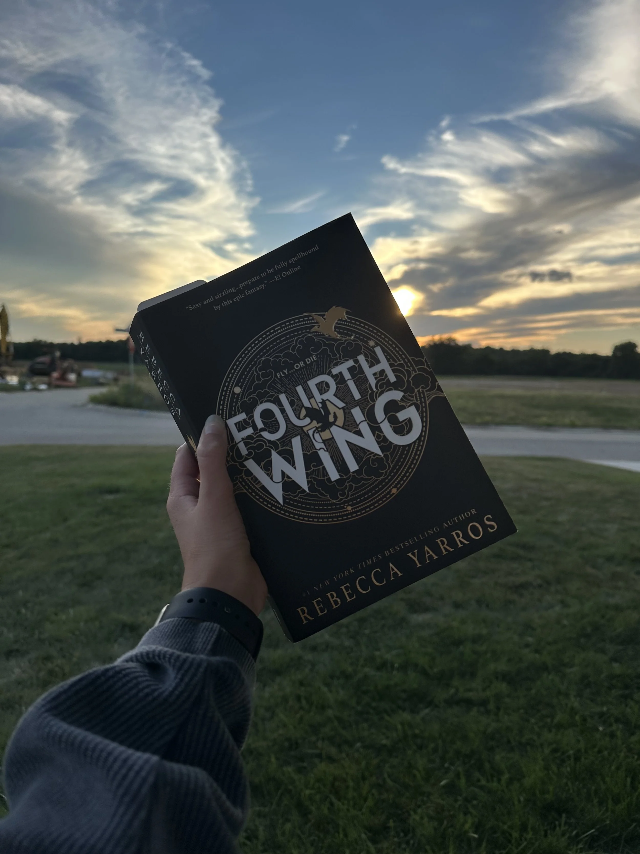 A person holding a book titled 'Fourth Wing' by Rebecca Yarros outdoors during sunset, with clouds and the sky in the background.