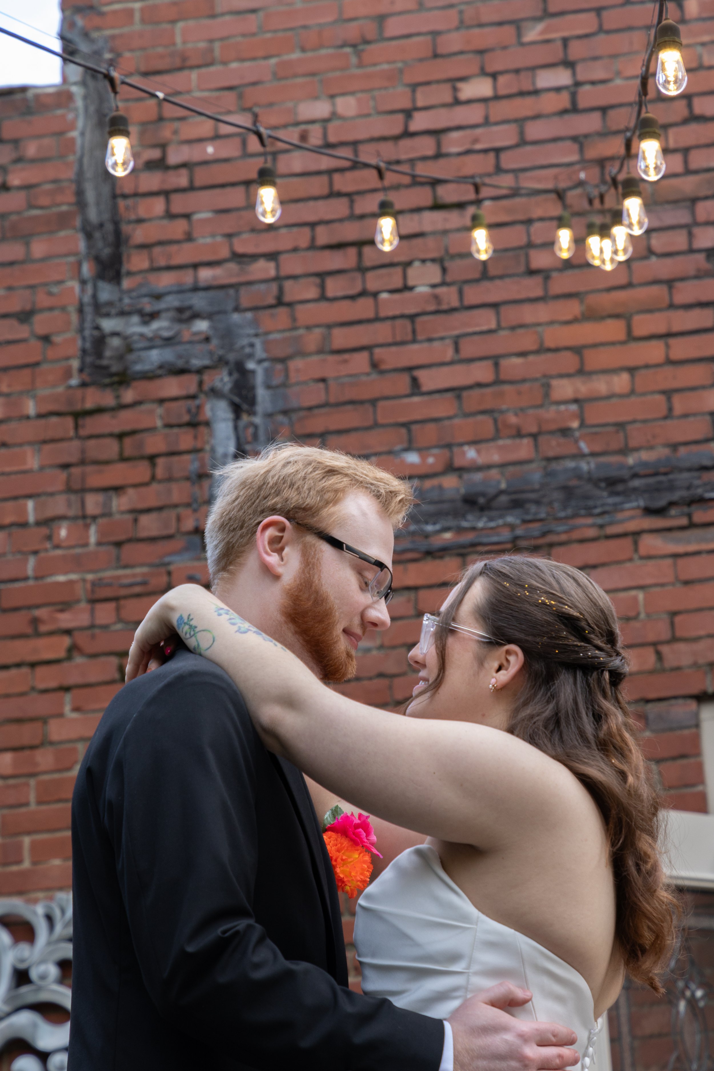 A couple in formal attire, a man with glasses and a woman with long hair, embracing and looking at each other in front of a brick wall with string lights hanging above them.