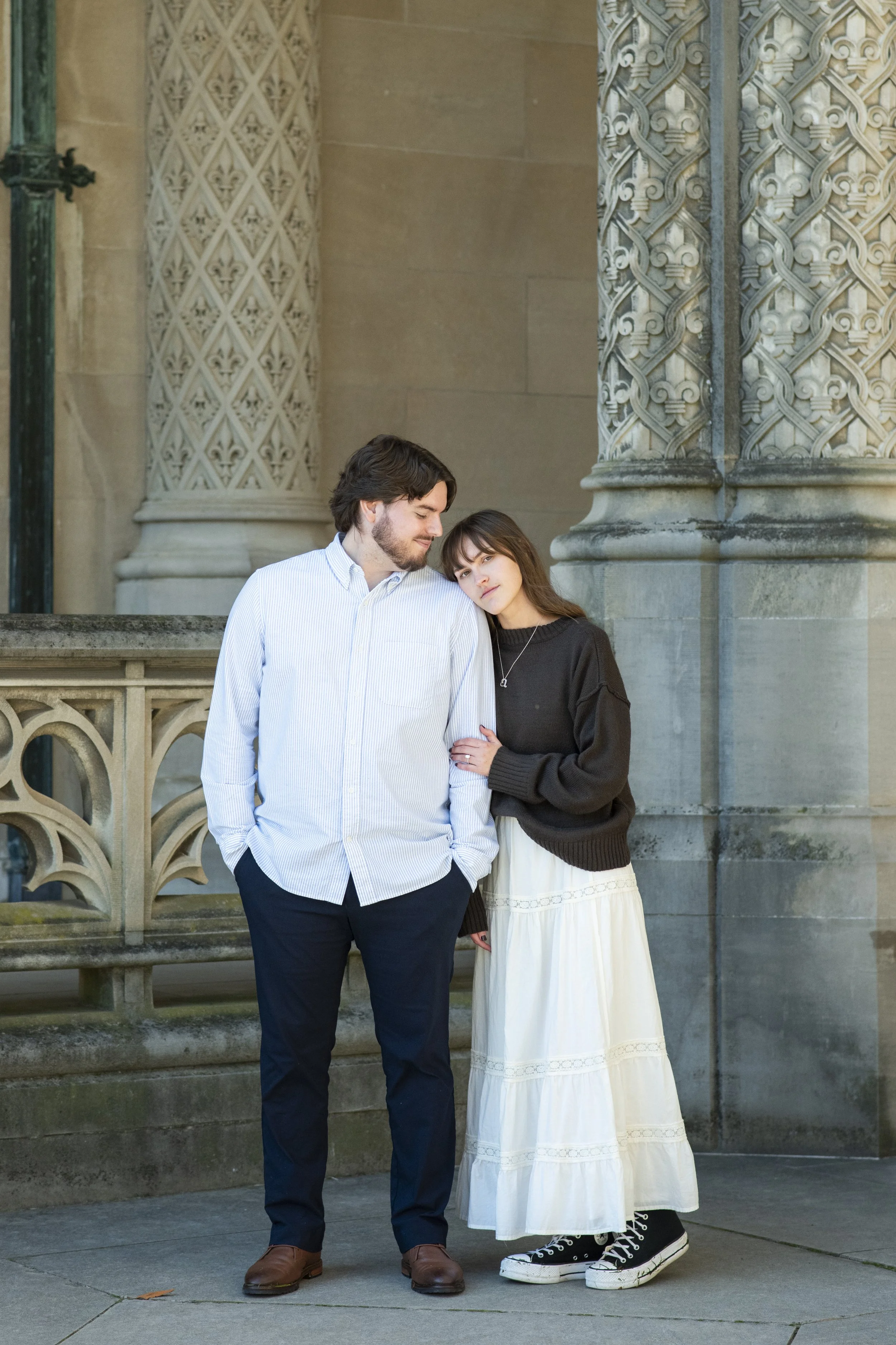 A man and woman standing close together on a sidewalk in front of an ornate stone building, with the woman leaning her head on the man's shoulder.