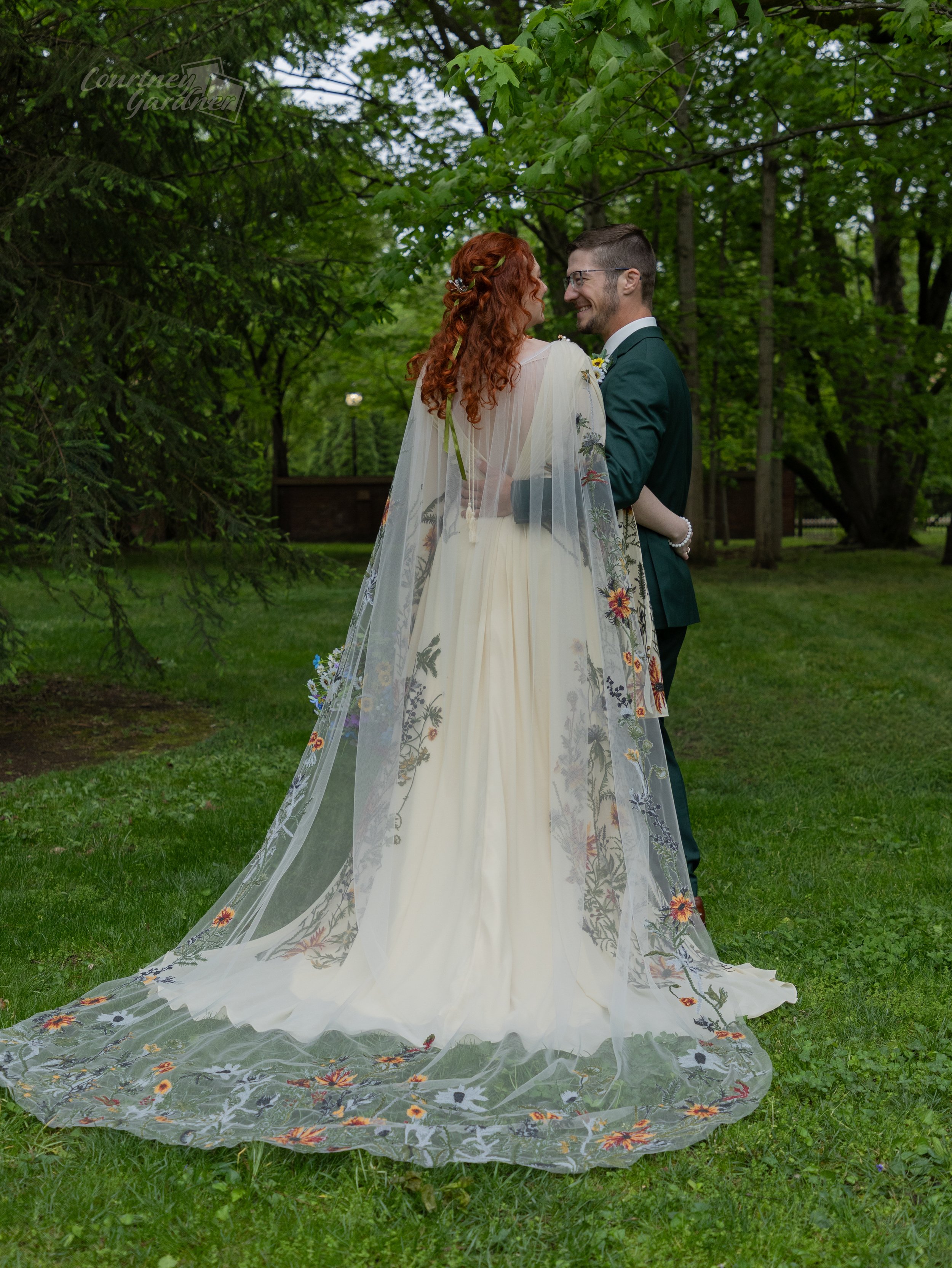 A bride and groom pose in a lush green park, facing each other and smiling. The bride has long, curly red hair and wears a light-colored dress with a sheer, embroidered cover-up, and a long train. The groom has short dark hair, glasses, and is dressed in a dark green suit with a white shirt.