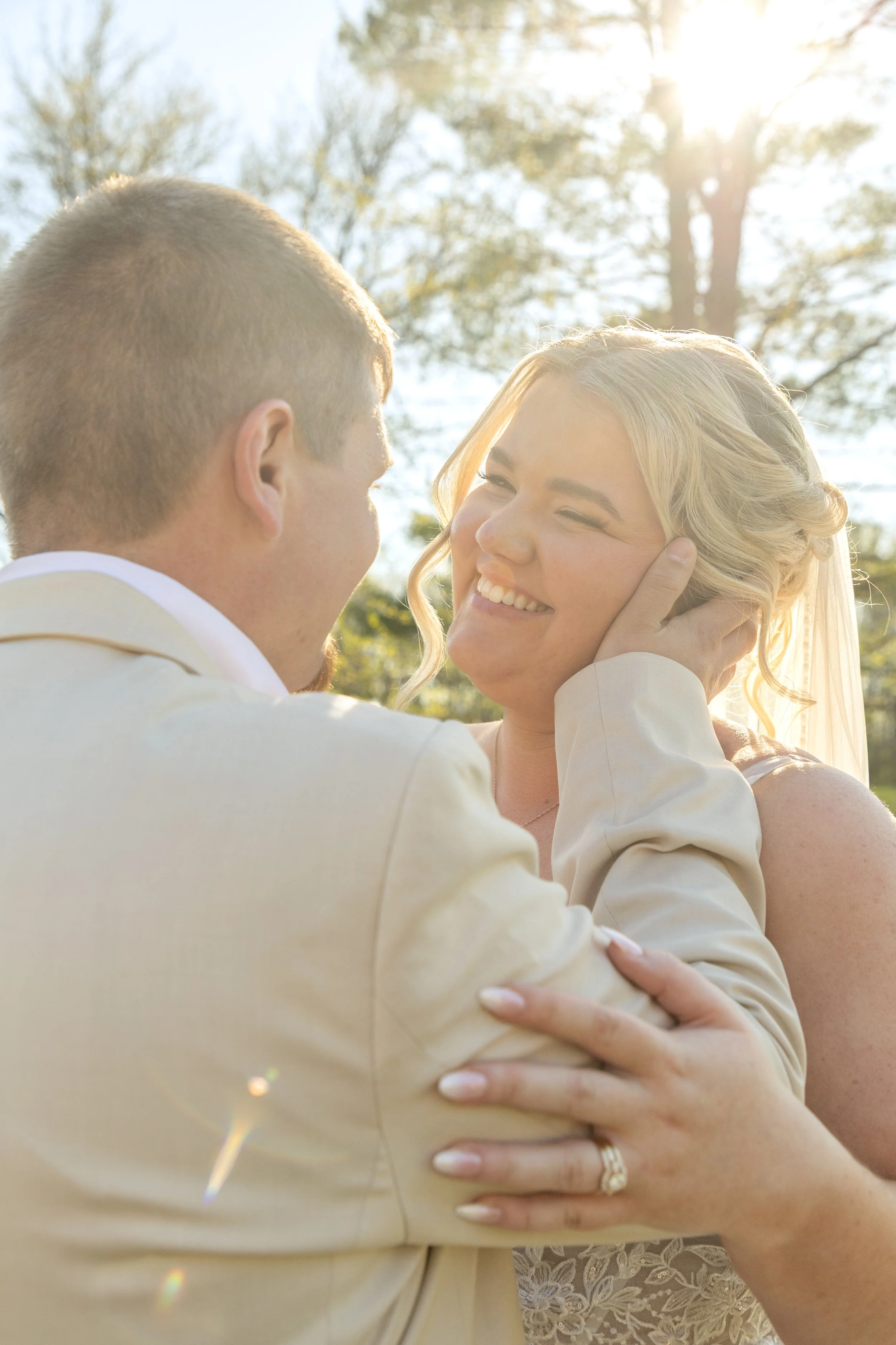 A bride and groom are seen in an intimate moment outdoors, with the groom holding the bride's face and smiling, under bright sunlight and trees in the background.