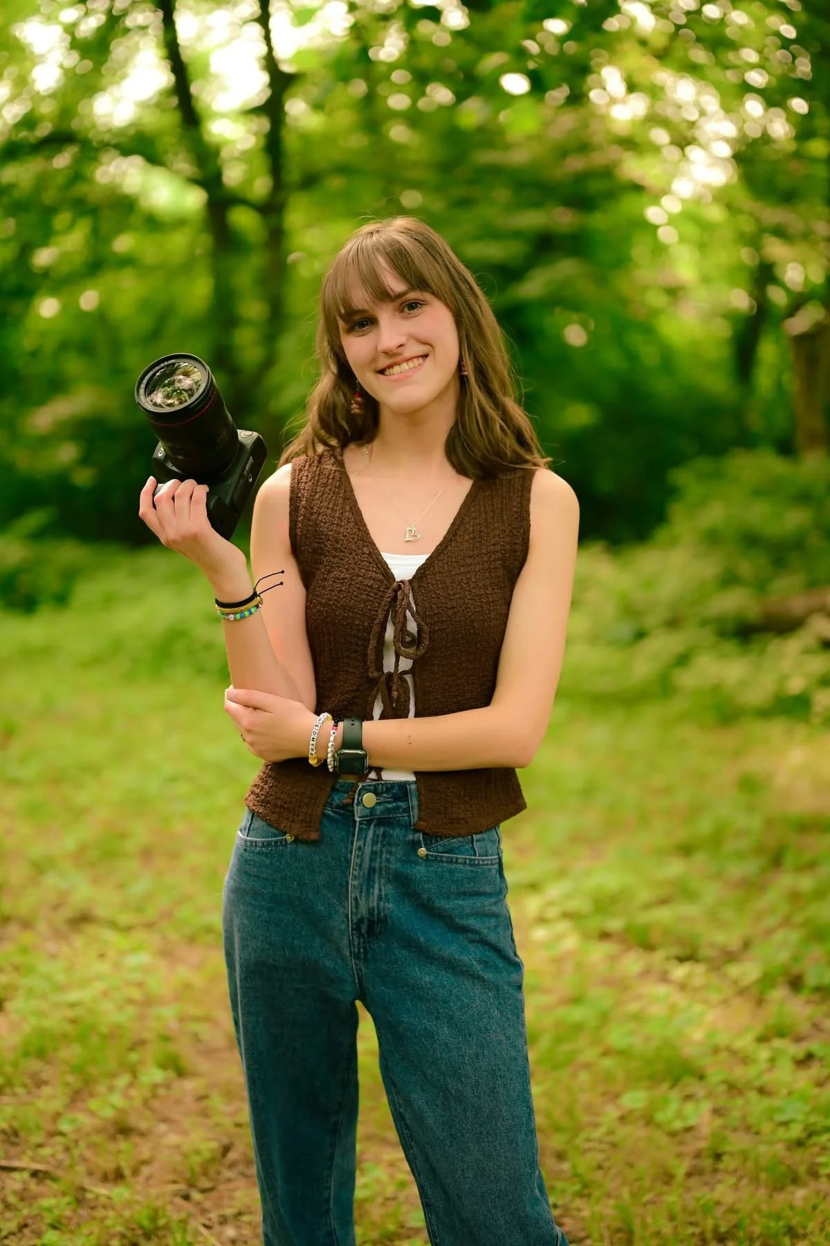 A young woman standing outdoors in a lush green forest, holding a camera camera in her right hand, smiling at the camera.