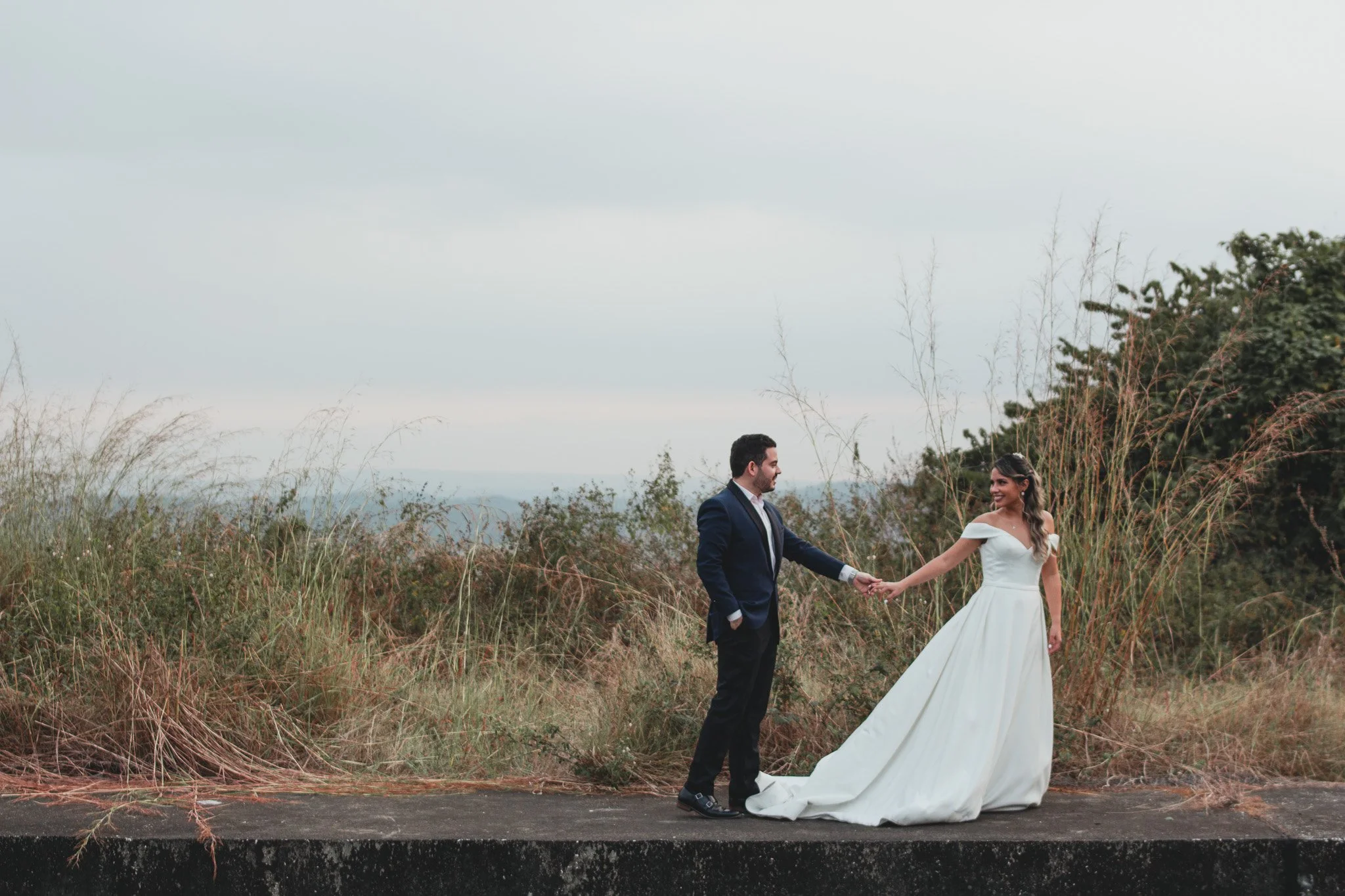 Una pareja de novios en un paisaje natural, ella con vestido de novia y él con traje oscuro, sosteniéndose de las manos.