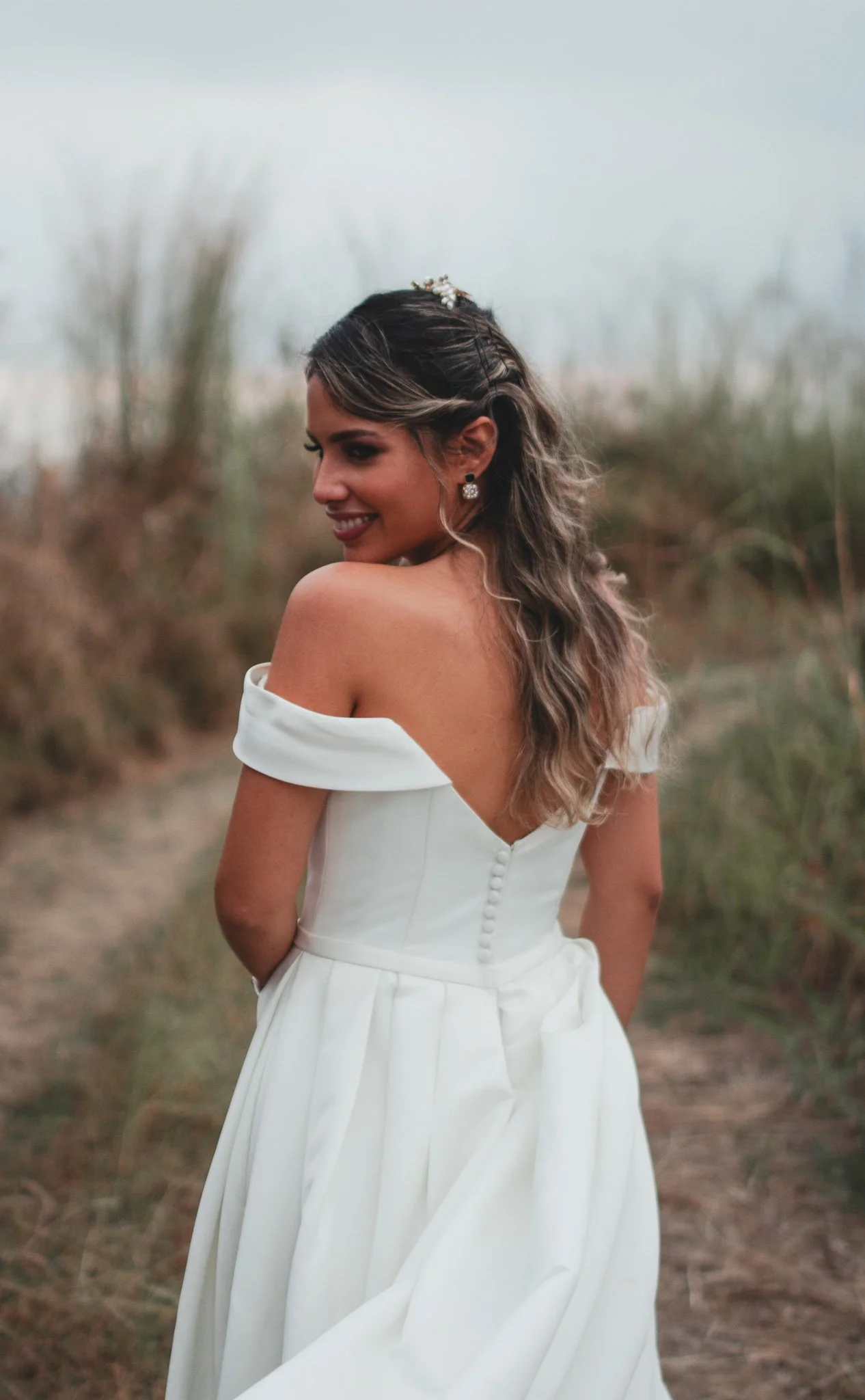 Mujer con vestido de novia blanco, de espaldas, en un camino rodeado de vegetación, sonriendo y mirando hacia atrás.