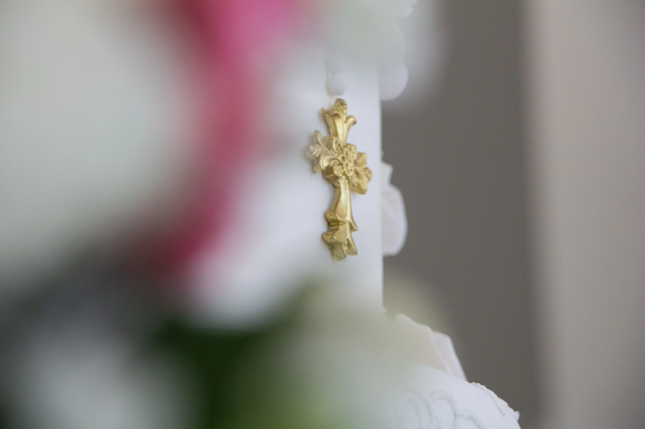 Detalle de una cruz dorada colgada en una pared blanca, con un fondo difuso que parece una figura de una estatua o escultura religiosa.