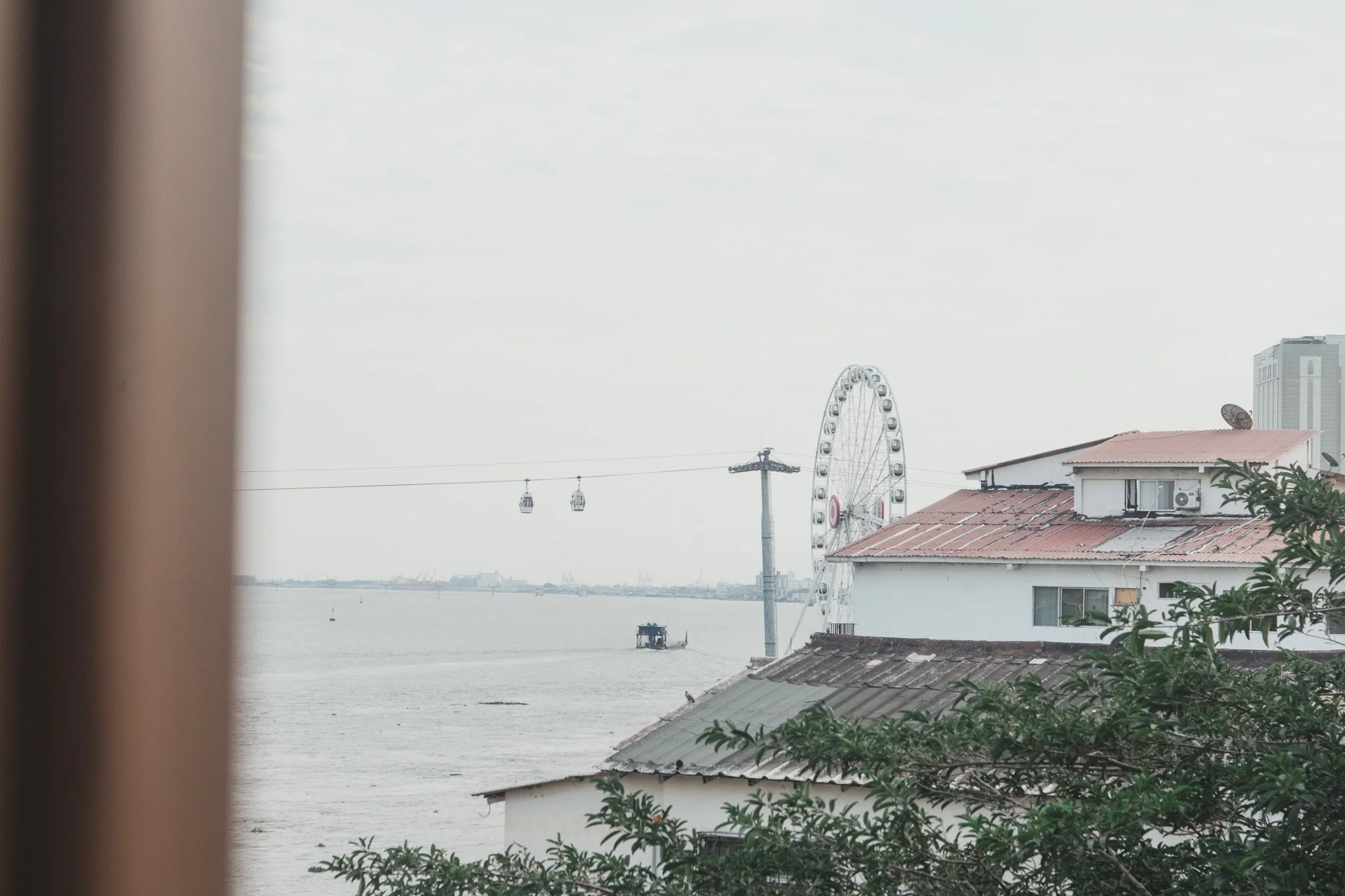 Vista de un río con una noria y un teleférico en el fondo, y casas con techos de lámina y vegetación en primer plano.