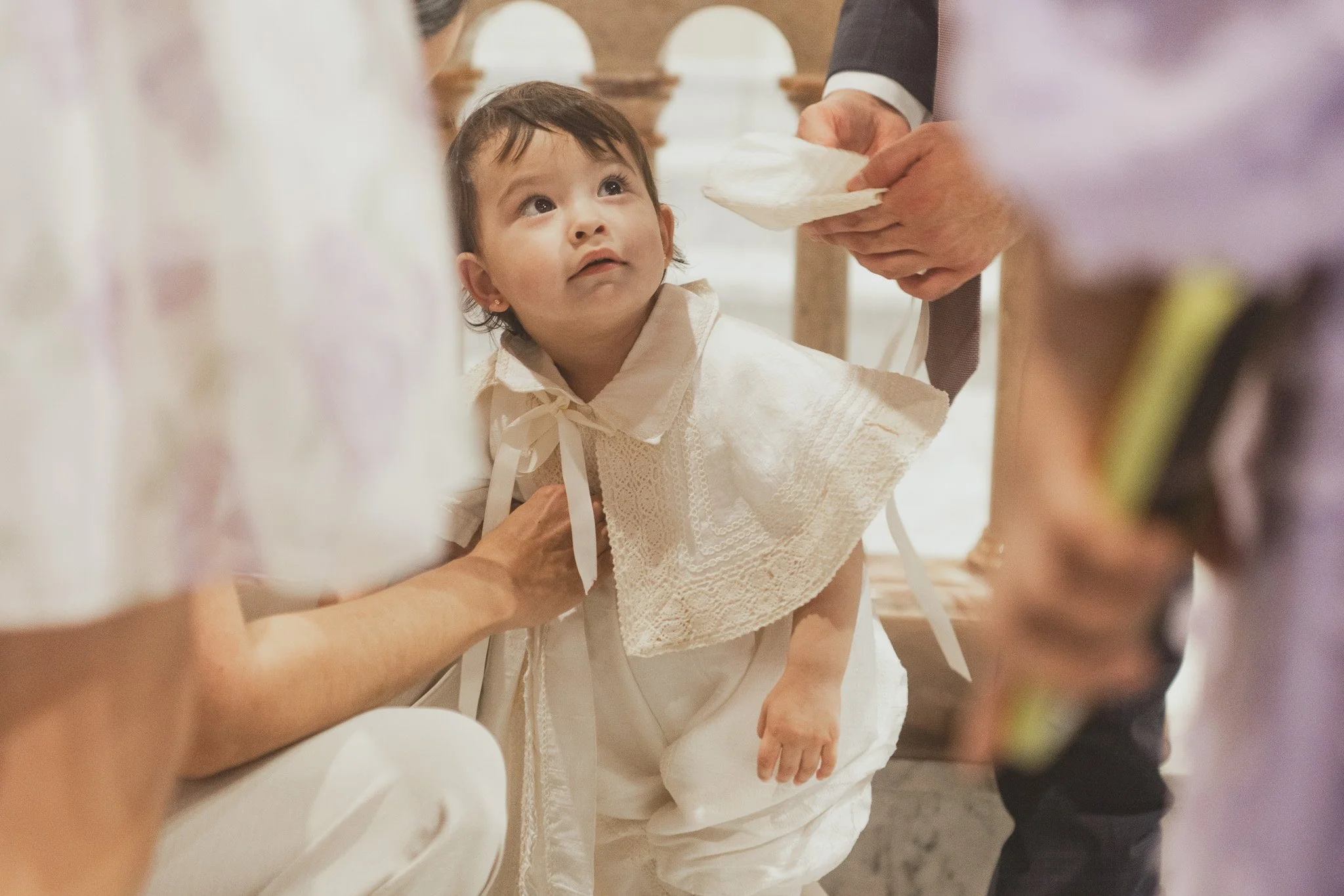 Niña en bautizo, con vestimenta blanca, en una ceremonia religiosa, rodeada de adultos que participan en la ceremonia en una iglesia.