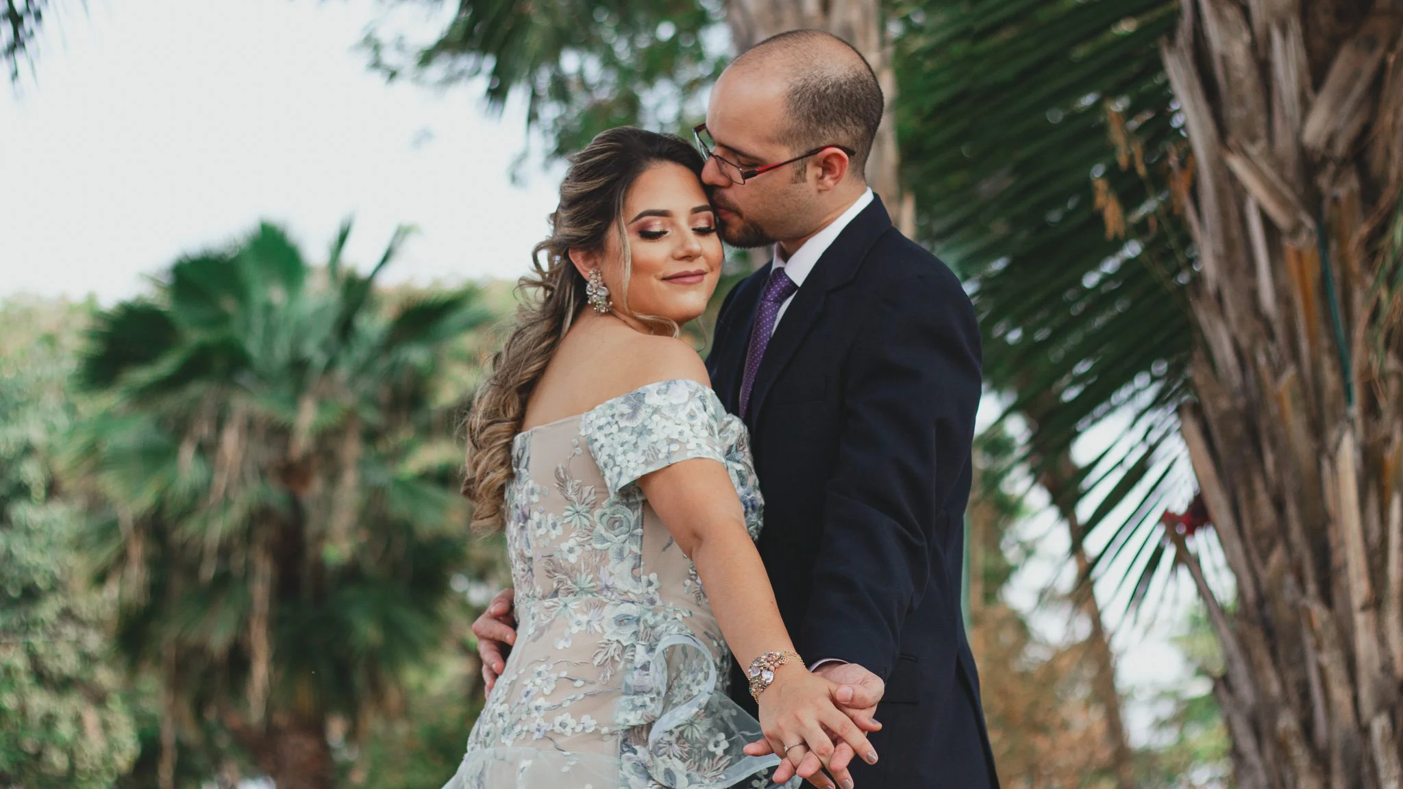 Una pareja de novios enamorados, la mujer con vestido de novia con encaje y el hombre con traje oscuro, en un entorno natural con árboles tropicales.