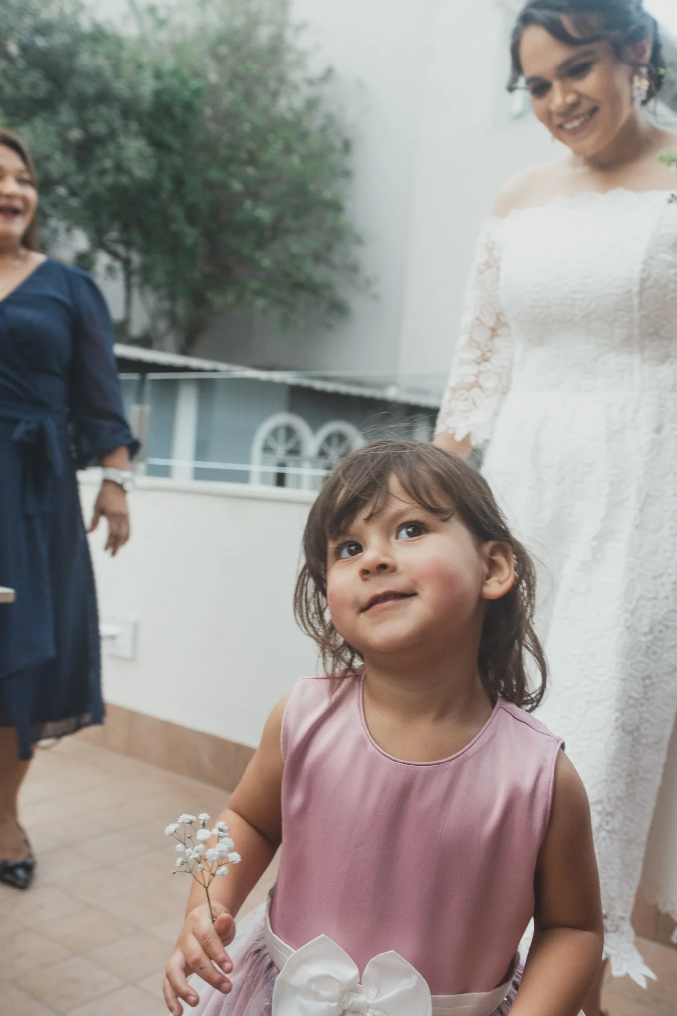 Una niña con vestido rosado y cinturón blanco, sosteniendo pequeñas flores blancas, en primer plano. Detrás, una mujer con vestido blanco y otra mujer con vestido azul, en un ambiente exterior con plantas verdes.