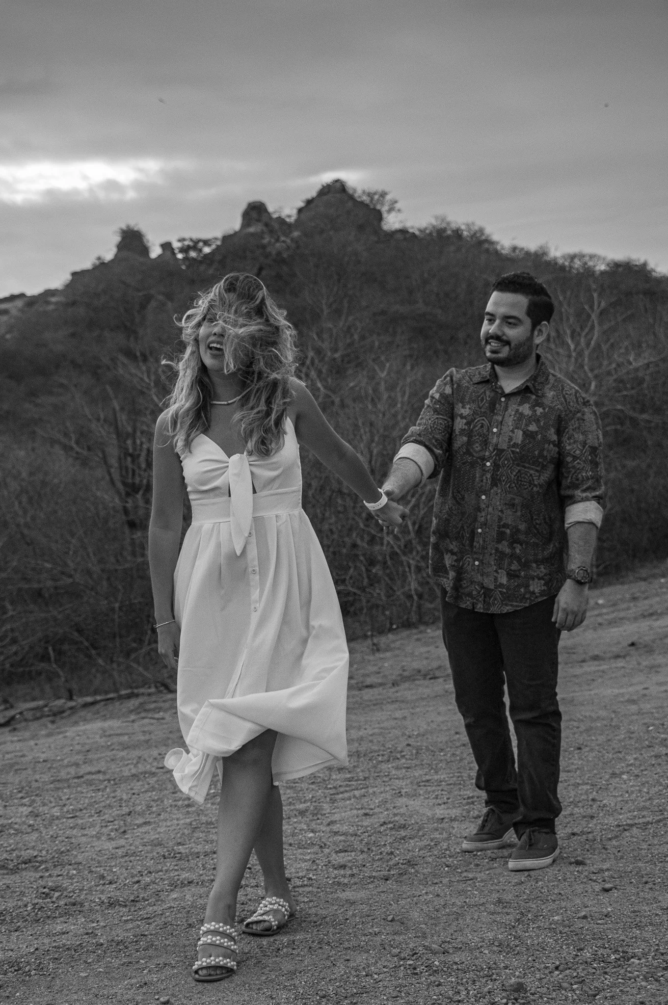 Una pareja caminando de la mano por un sendero al aire libre con montañas y árboles al fondo, en un día nublado.