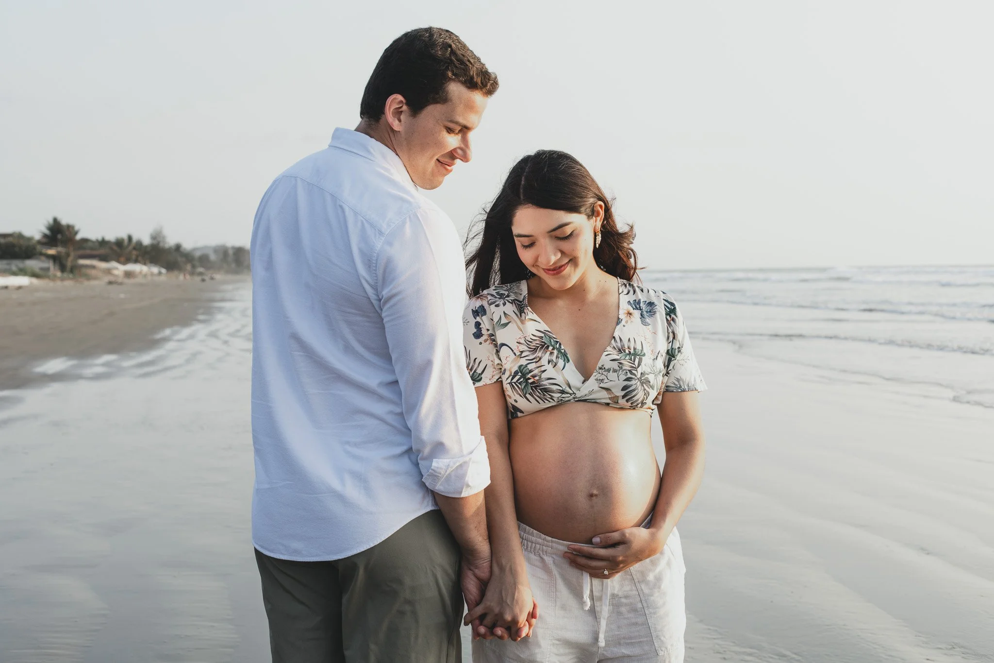 Una pareja en la playa, ella embarazada, se toman de la mano y miran con cariño, mientras la mujer sostiene su vientre.