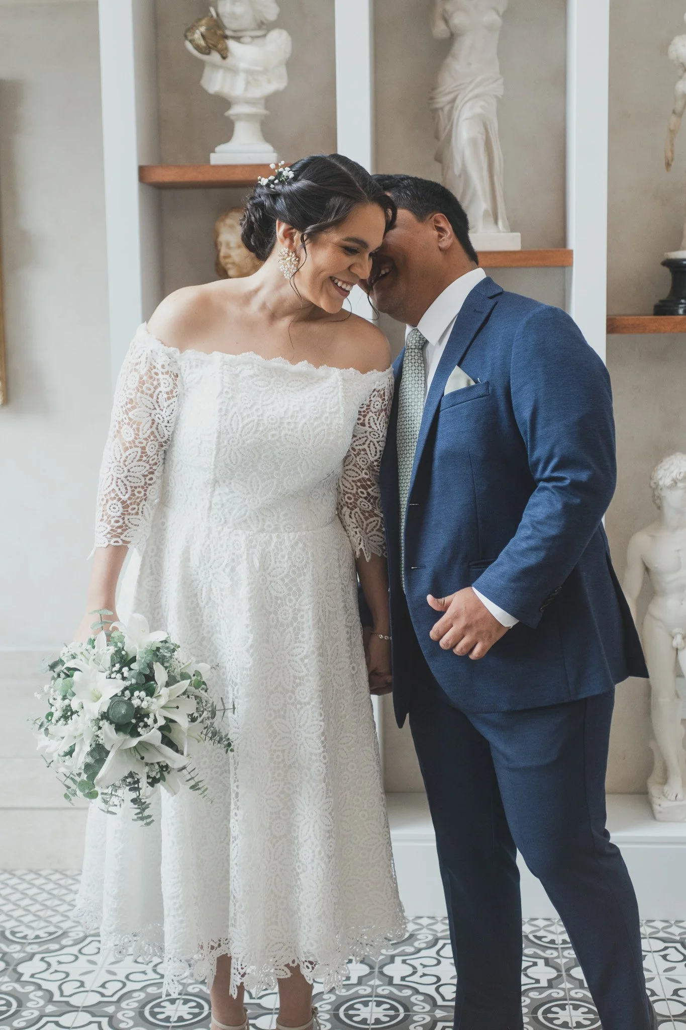 Pareja de novios en su boda, acercándose y sonriendo, con fondos de estatuas clásicas, la novia sostiene un ramo de flores blancas.