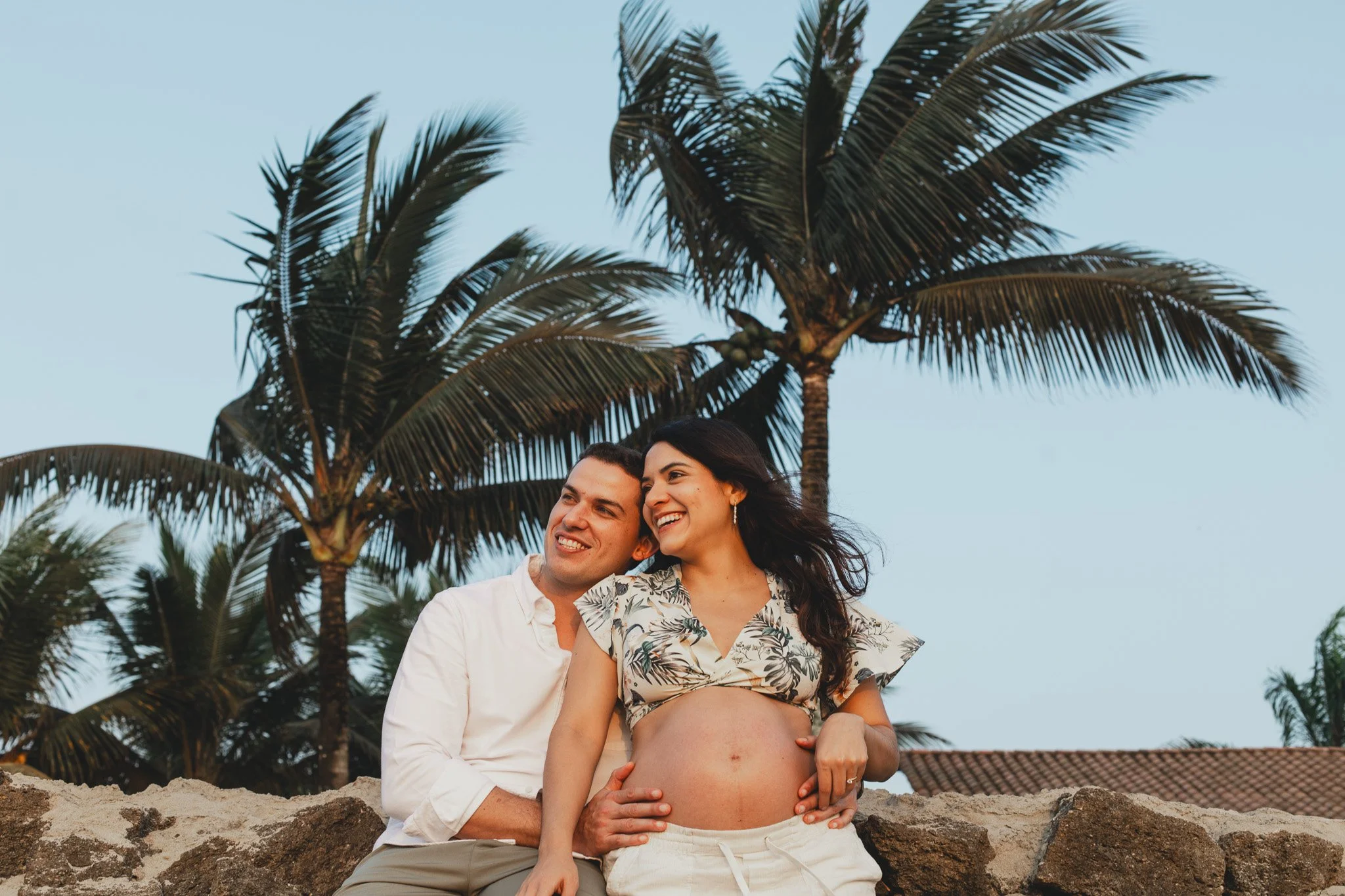 Una mujer embarazada y un hombre sonrientes sentados en una pared de piedra junto a palmeras, en un escenario tropical al atardecer.