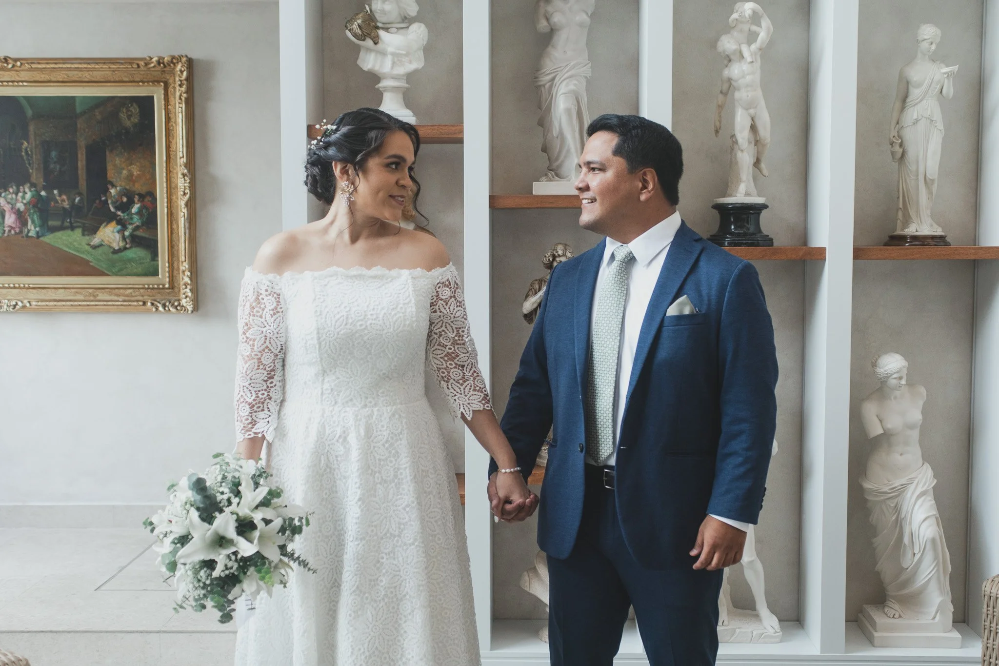 Pareja de novios en ceremonia de bodas, la mujer con vestido blanco y ramo, el hombre con traje azul, en interior decorado con esculturas y un cuadro