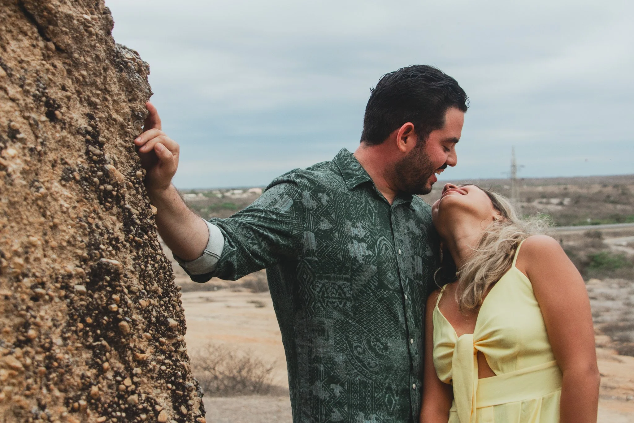 Una pareja en una escarpada zona rocosa, ella de vestido amarillo y él con camisa de estampado oscuro, se miran y sonríen con cariño, la escena expresa amor y alegría en un paisaje árido.