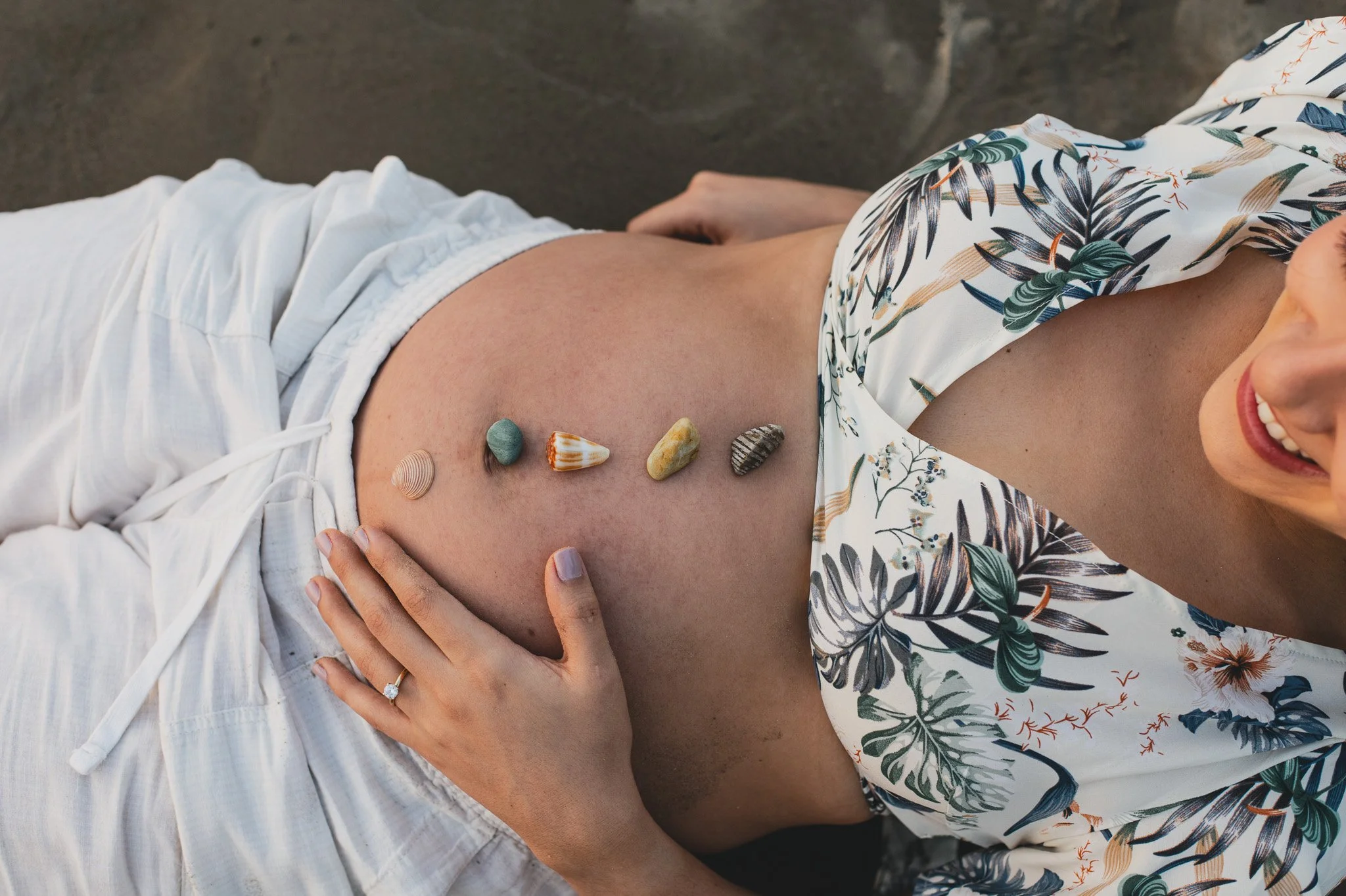 Mujer en vestido con estampado floral acostada, con piedras y conchas sobre su abdomen desnudo, sonriendo