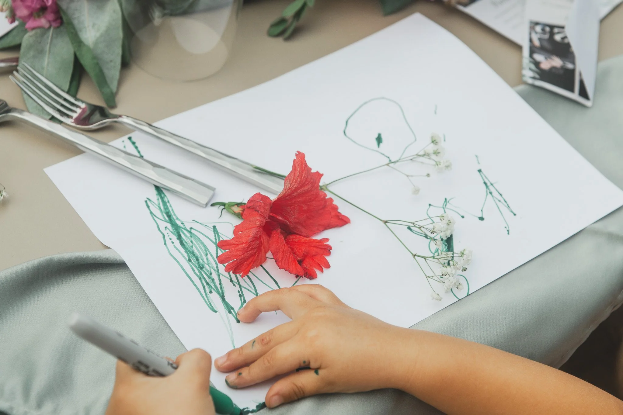 Mano dibuja en papel con flor roja y pequeñas flores blancas, utensilios de mesa sobre la mesa. Hay hojas verdes y otros objetos alrededor.
