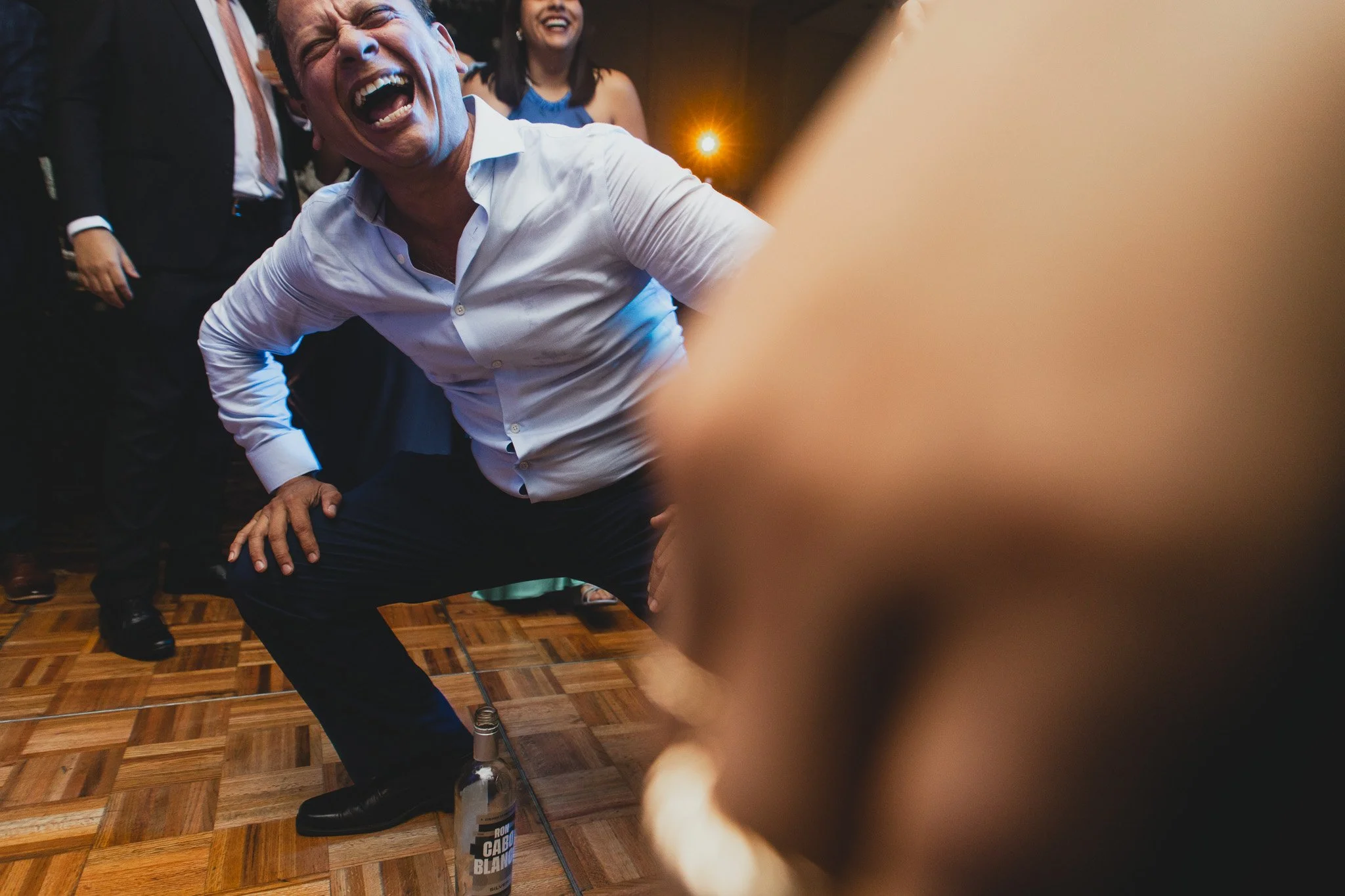 Hombre riendo y bailando en fiesta, con botella en el suelo y otras personas de fondo.