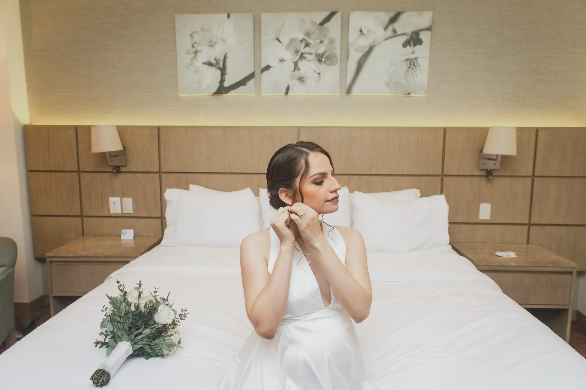 Mujer vestida de novia arreglándose en una habitación con cama de sábanas blancas, con una bola de flores en la cama, en una habitación moderna con cuadros de flores en la pared.