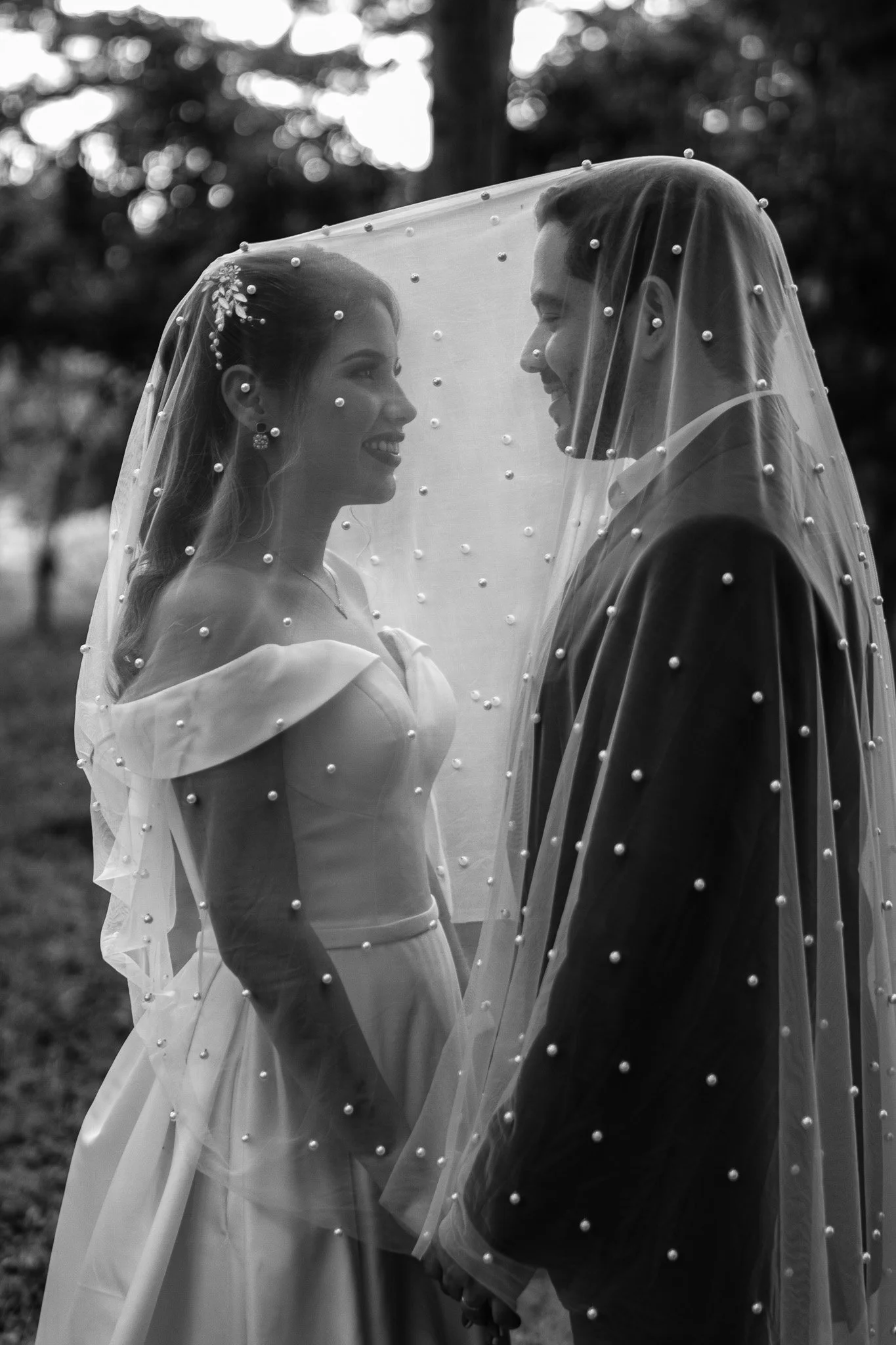 Una pareja de novios con velo de novia, sosteniéndose de las manos y sonriendo frente a frente en un entorno exterior, en blanco y negro.