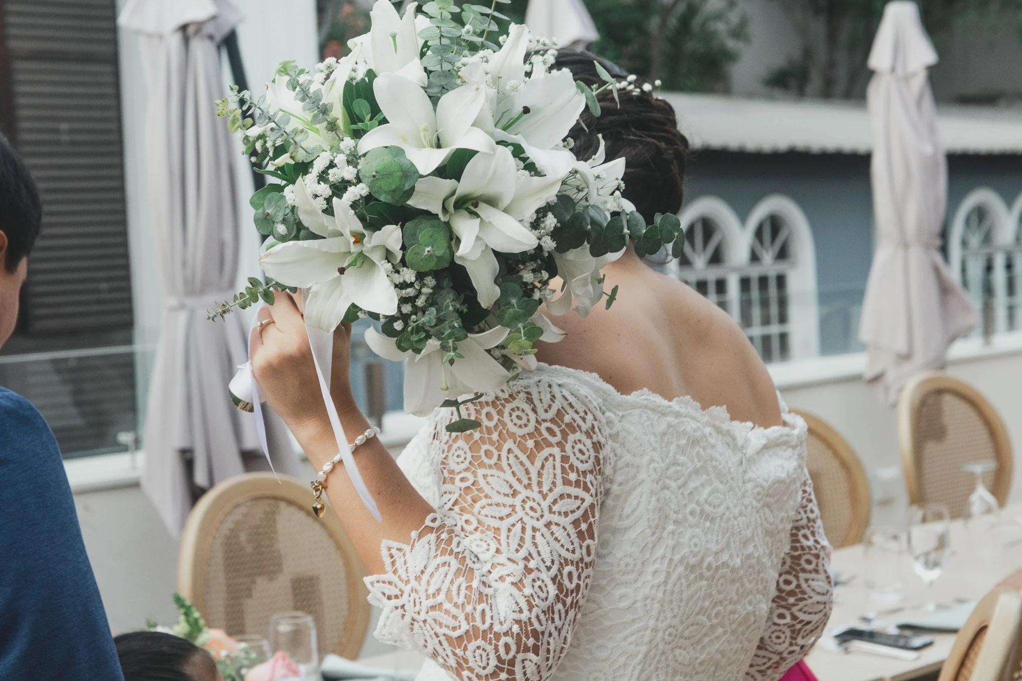 Mujer con vestido de encaje blanco y un ramo de flores grandes y blancas en rostro en una celebración al aire libre.