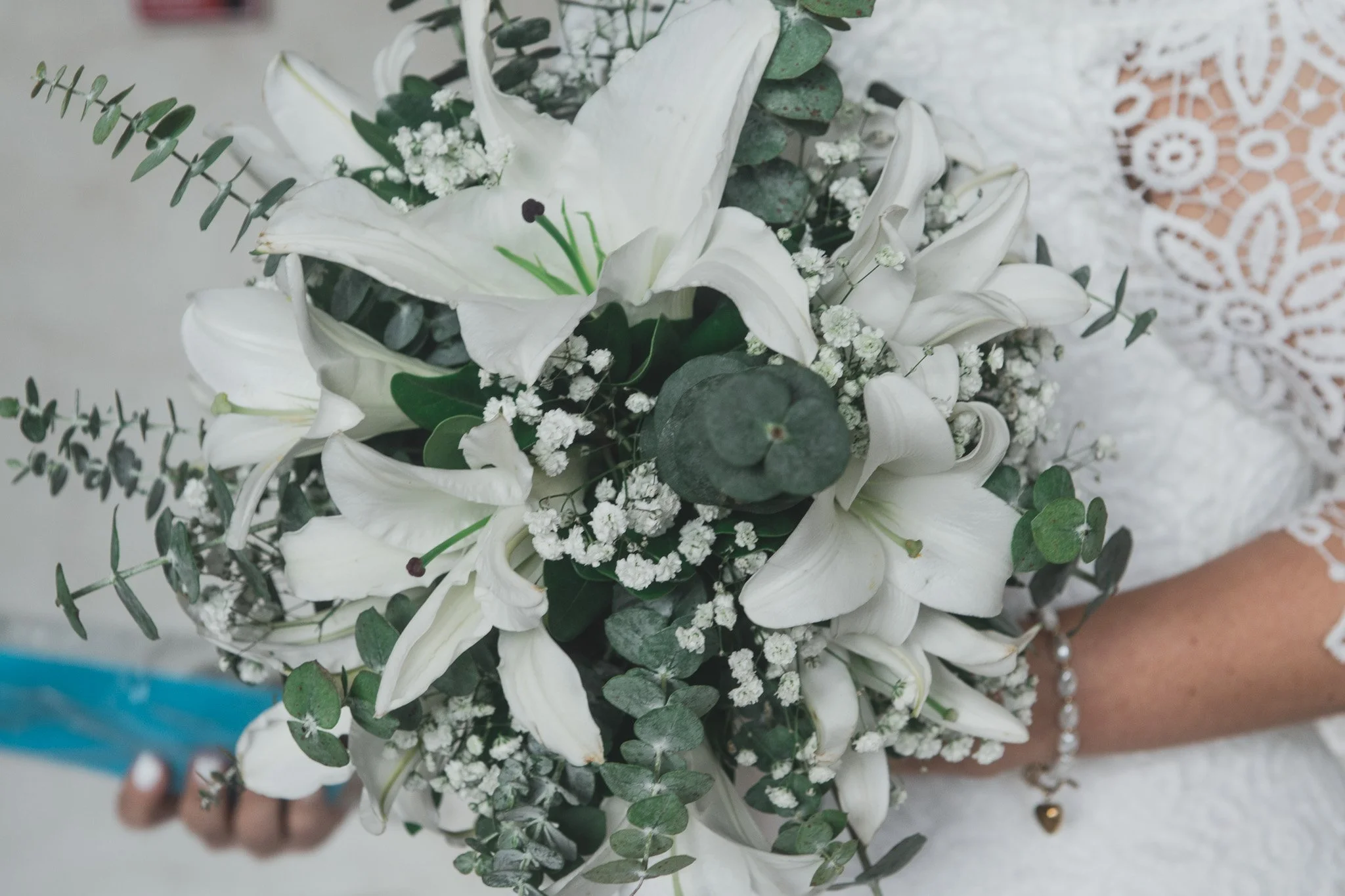 Ramo de flores blancas con lirios, eucalipto y flores pequeñas, sostenido por una persona con un vestido de encaje blanco.