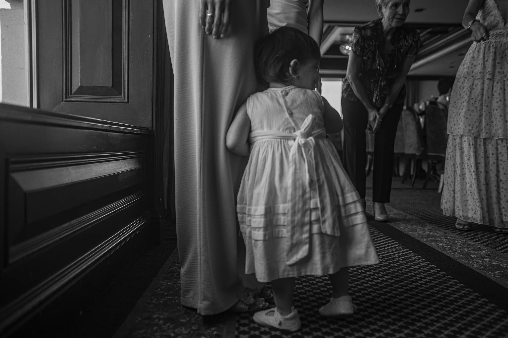 Niña en vestido esperando detrás de una pared en un evento social, con otras personas adultas en el fondo.