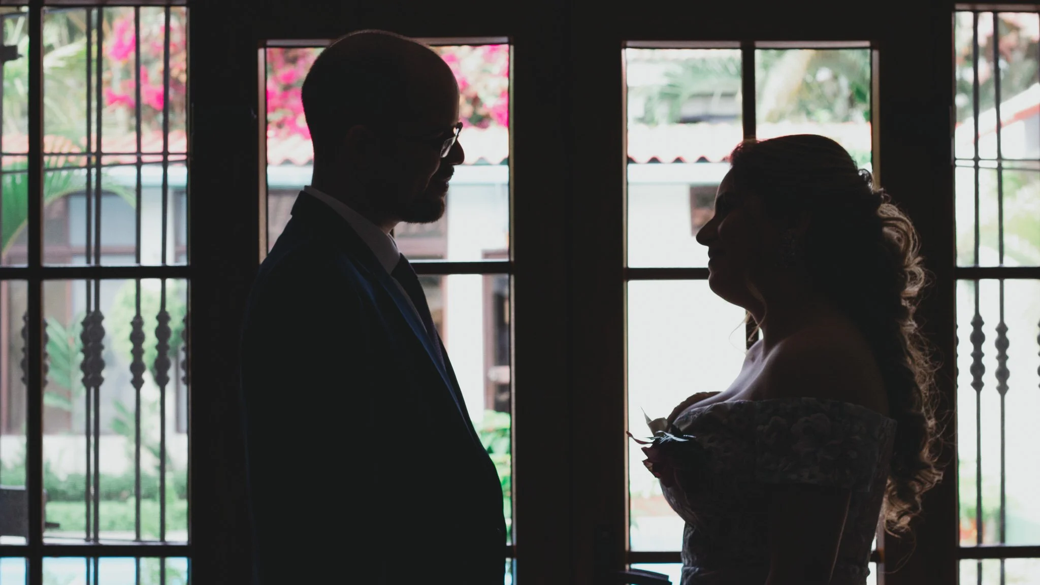 Pareja en silueta frente a una ventana con rejas de metal, la mujer lleva vestido de novia y el hombre un traje de novia, en un ambiente interior.