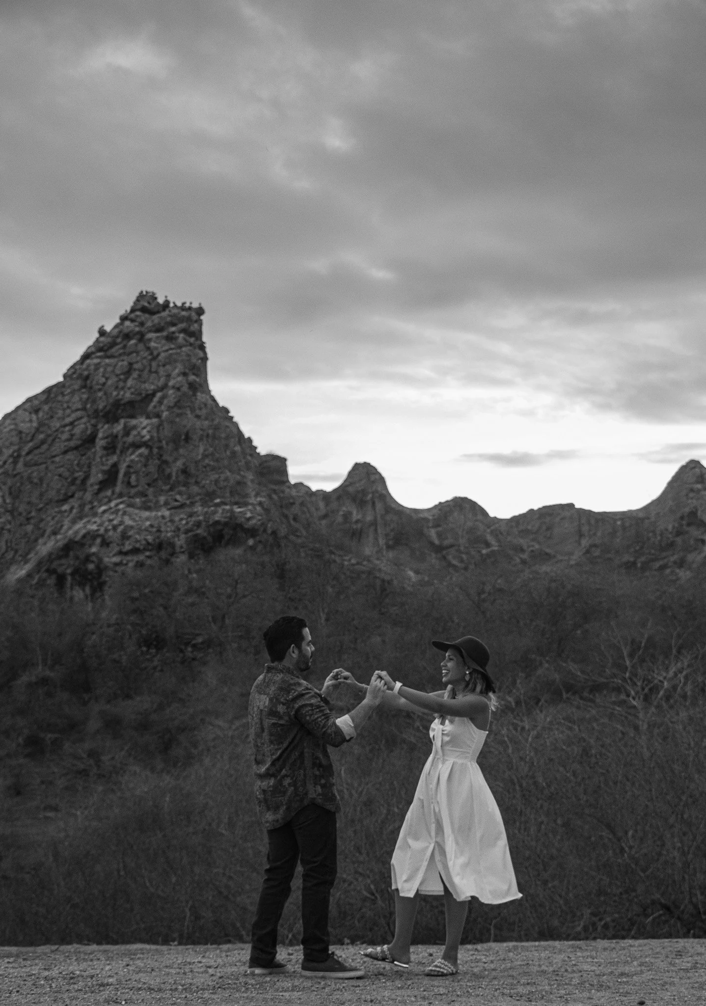 Una pareja de espaldas, un hombre y una mujer, sosteniéndose de las manos en un paisaje con montañas y cielo nublado en blanco y negro.