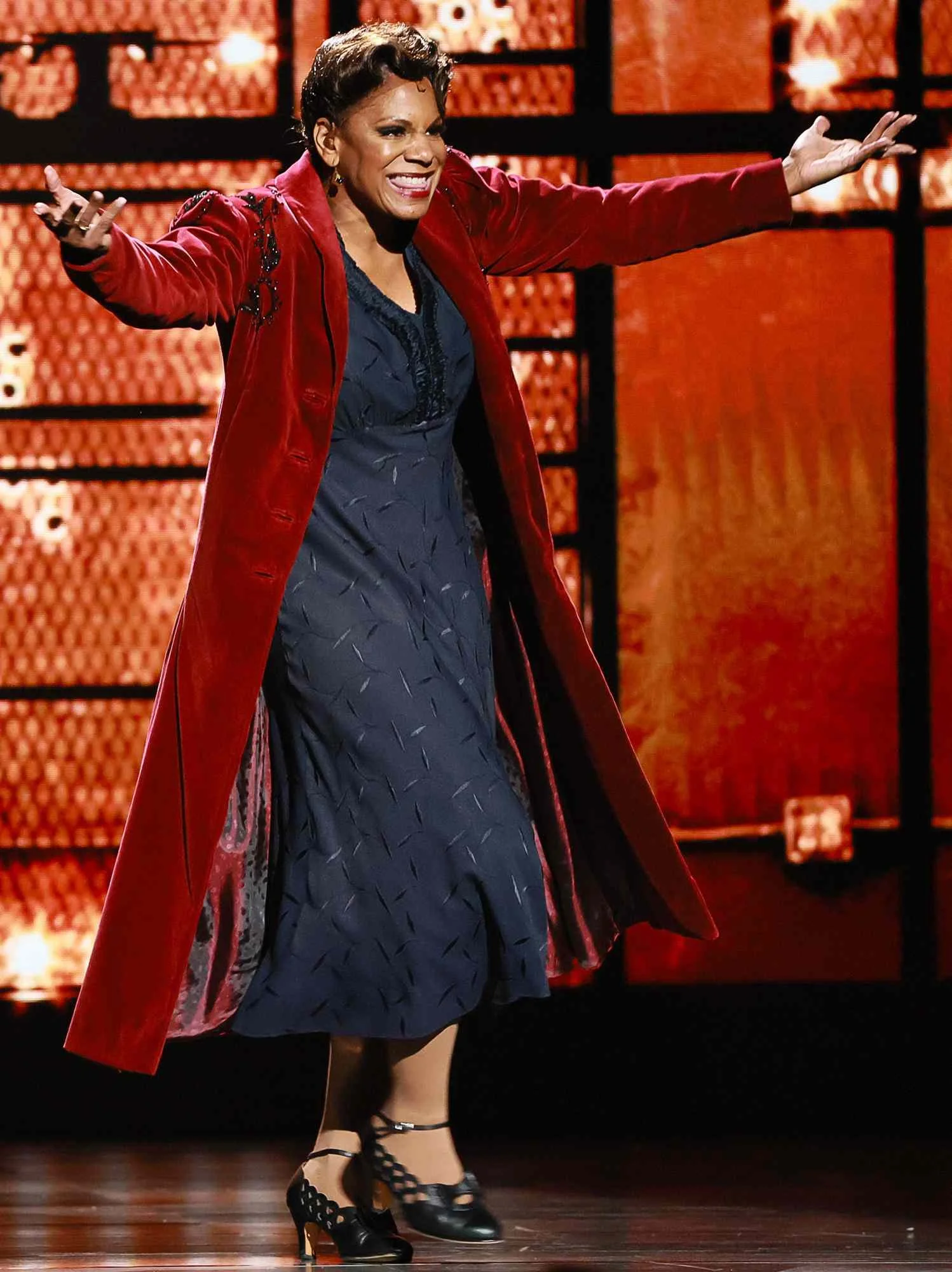Woman with short hair wearing a red coat over a dark dress, dancing on stage with arms outstretched, a warm orange background.