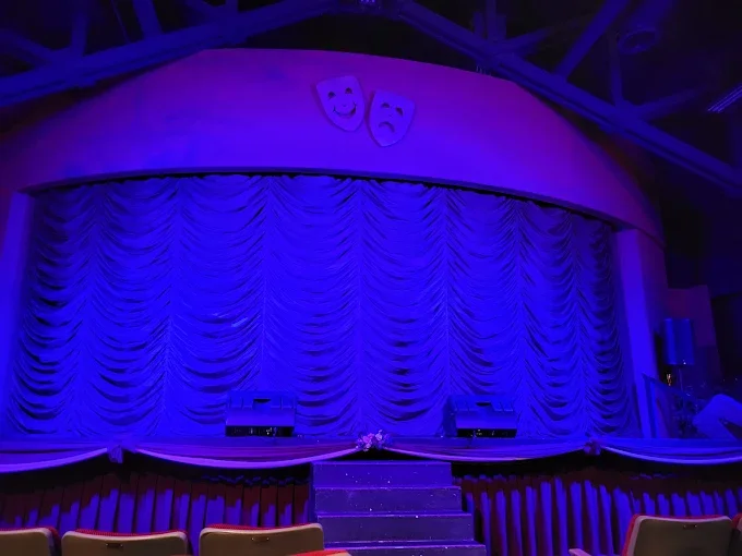 The theater stage is set with blue lighting, featuring a curtain backdrop and two comedy and tragedy masks hanging above it.