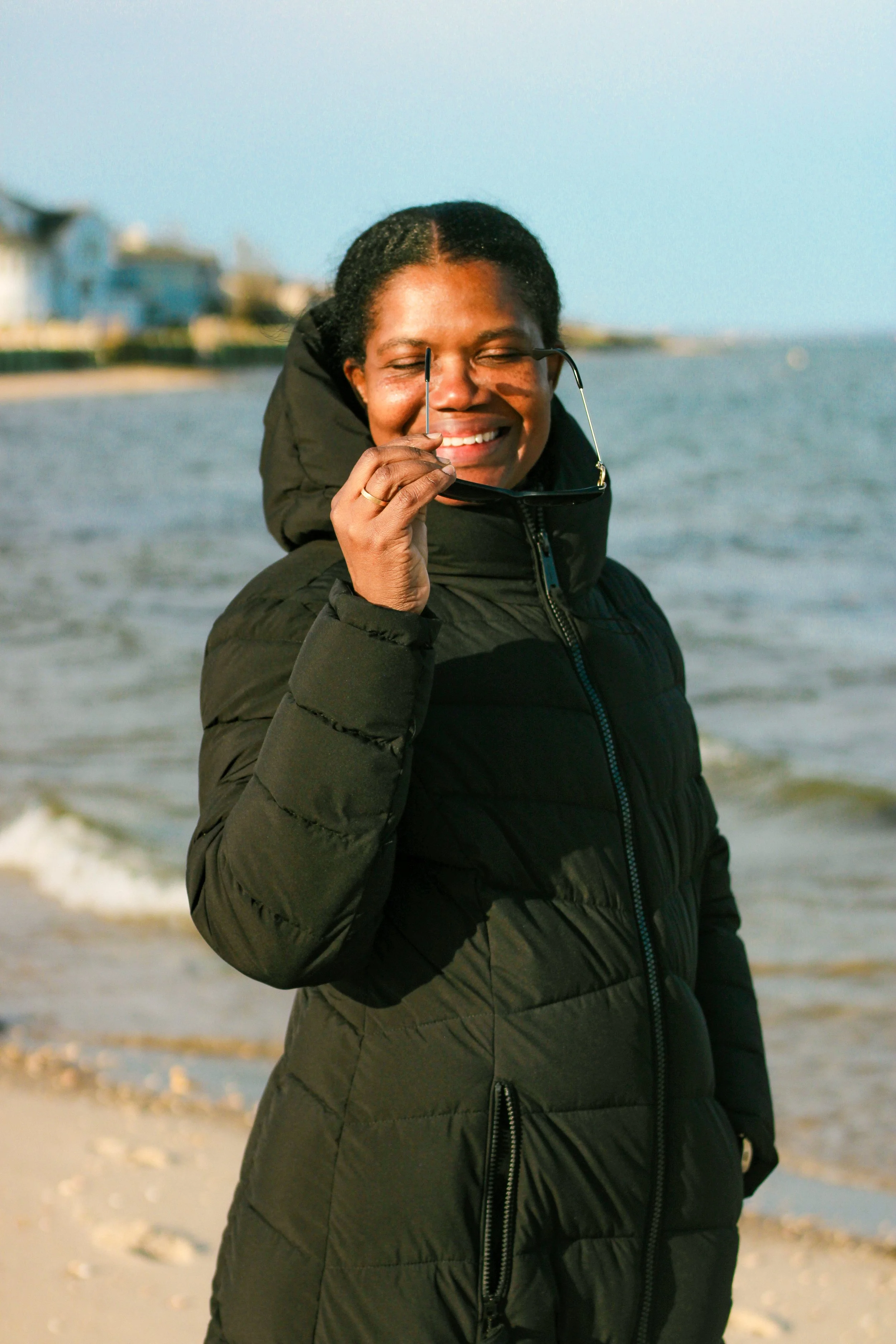 A woman outdoors by the water, wearing a black puffer jacket, smiling, holding sunglasses, with a shoreline and buildings in the background on a sunny day.