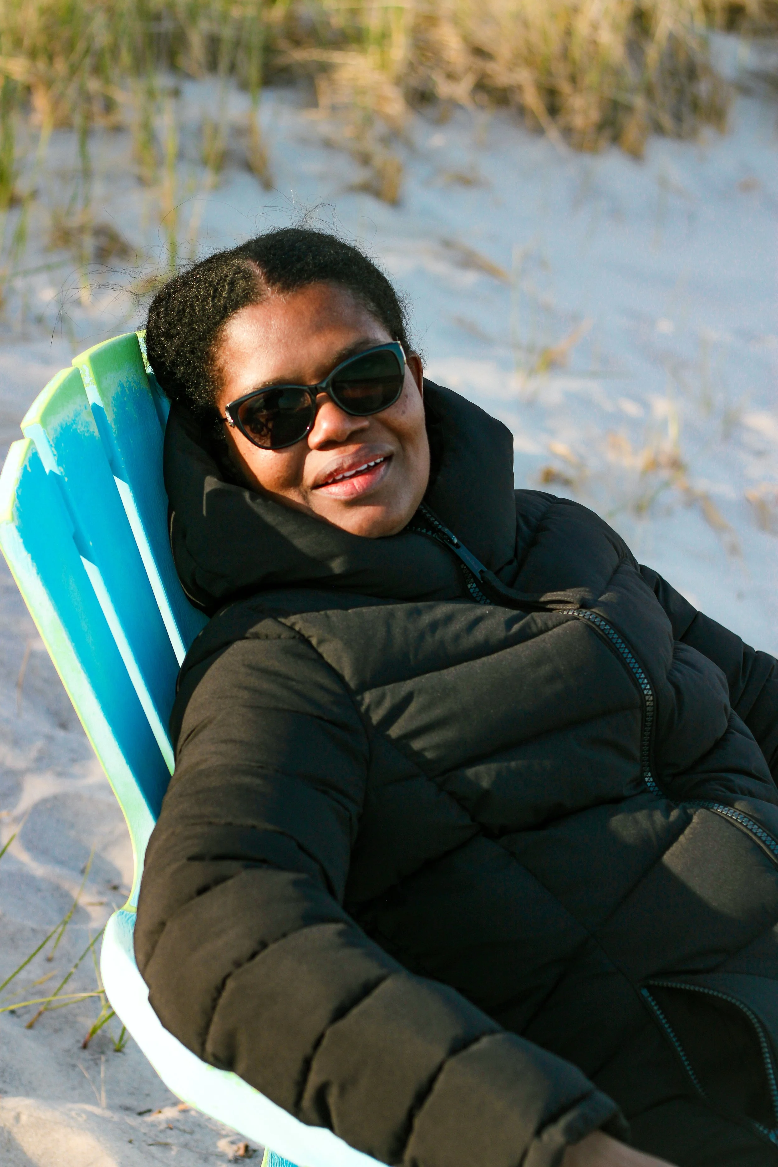 Woman sitting on a beach chair on sandy beach with grass in the background, wearing sunglasses and a black jacket, smiling.