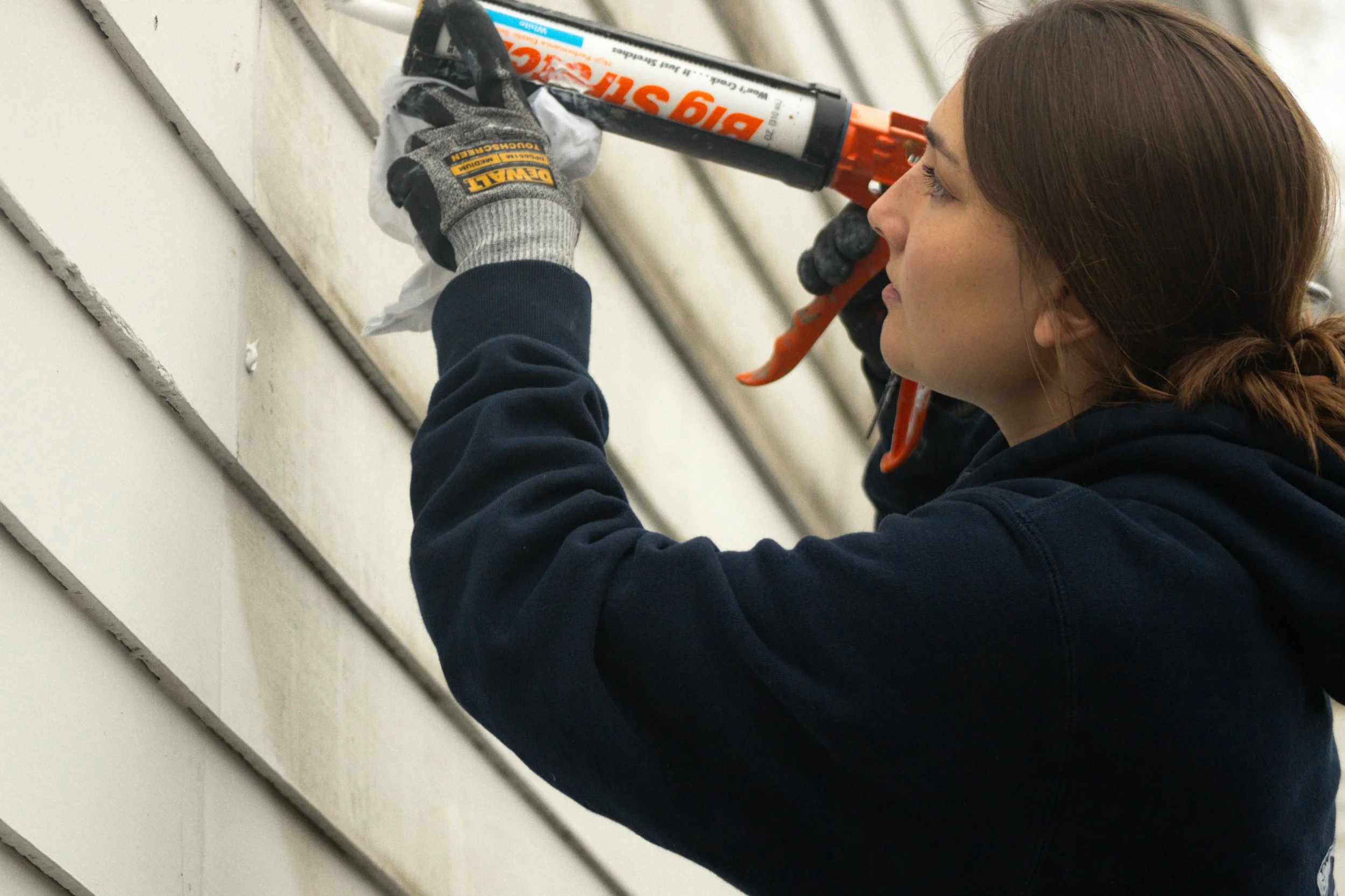 A woman with brown hair wearing a black hoodie and gray gloves applying white exterior paint to house siding with a caulking gun.