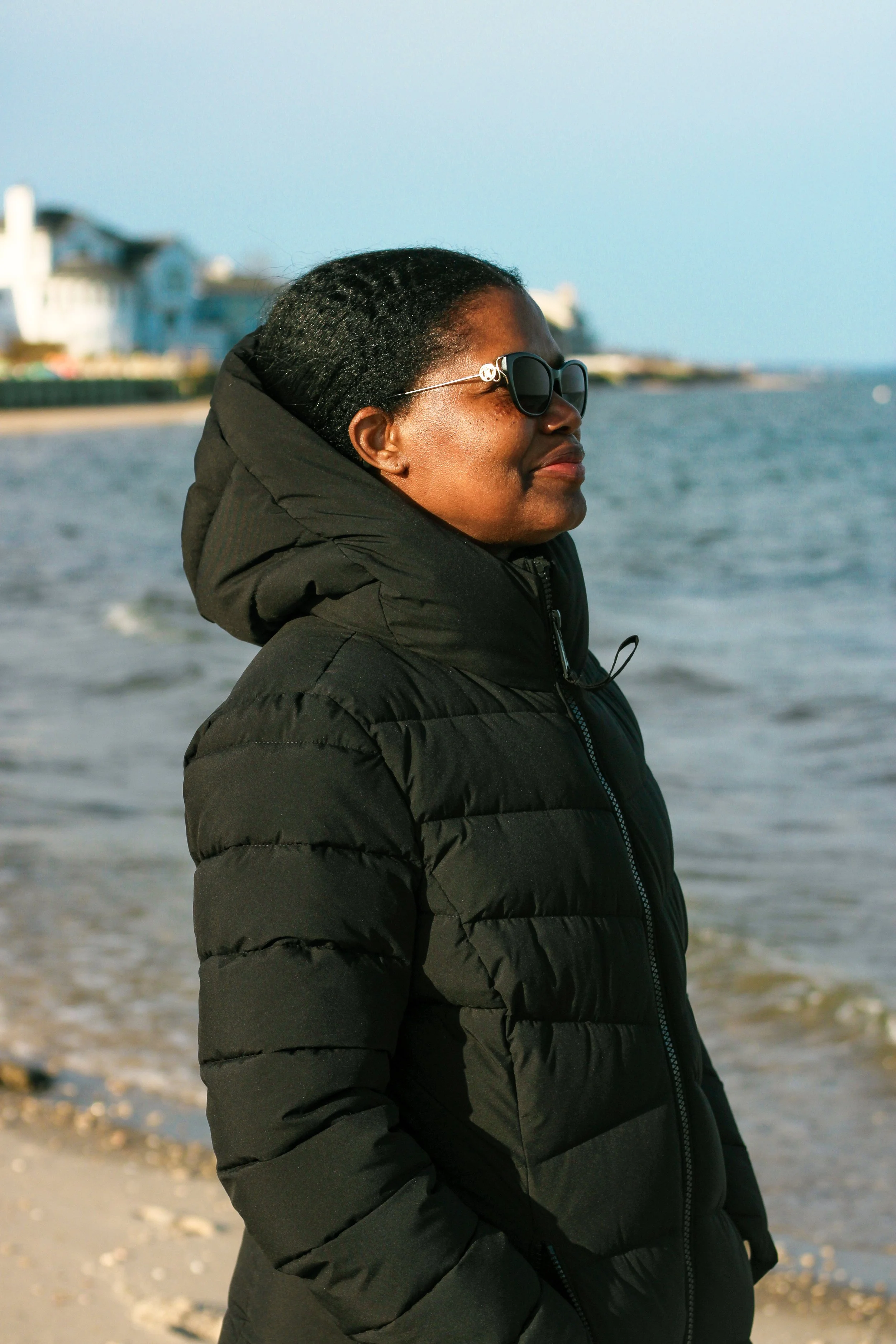 A woman wearing a black puffy jacket and sunglasses standing on a beach with ocean waves in the background.