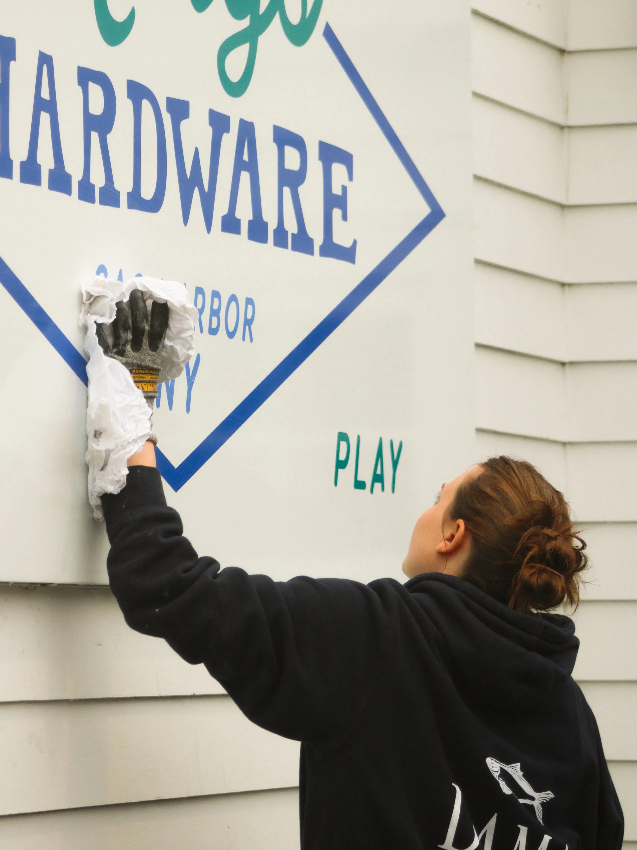 Young woman putting up a sign on a light-colored house wall, wearing a black hoodie with a fish design.