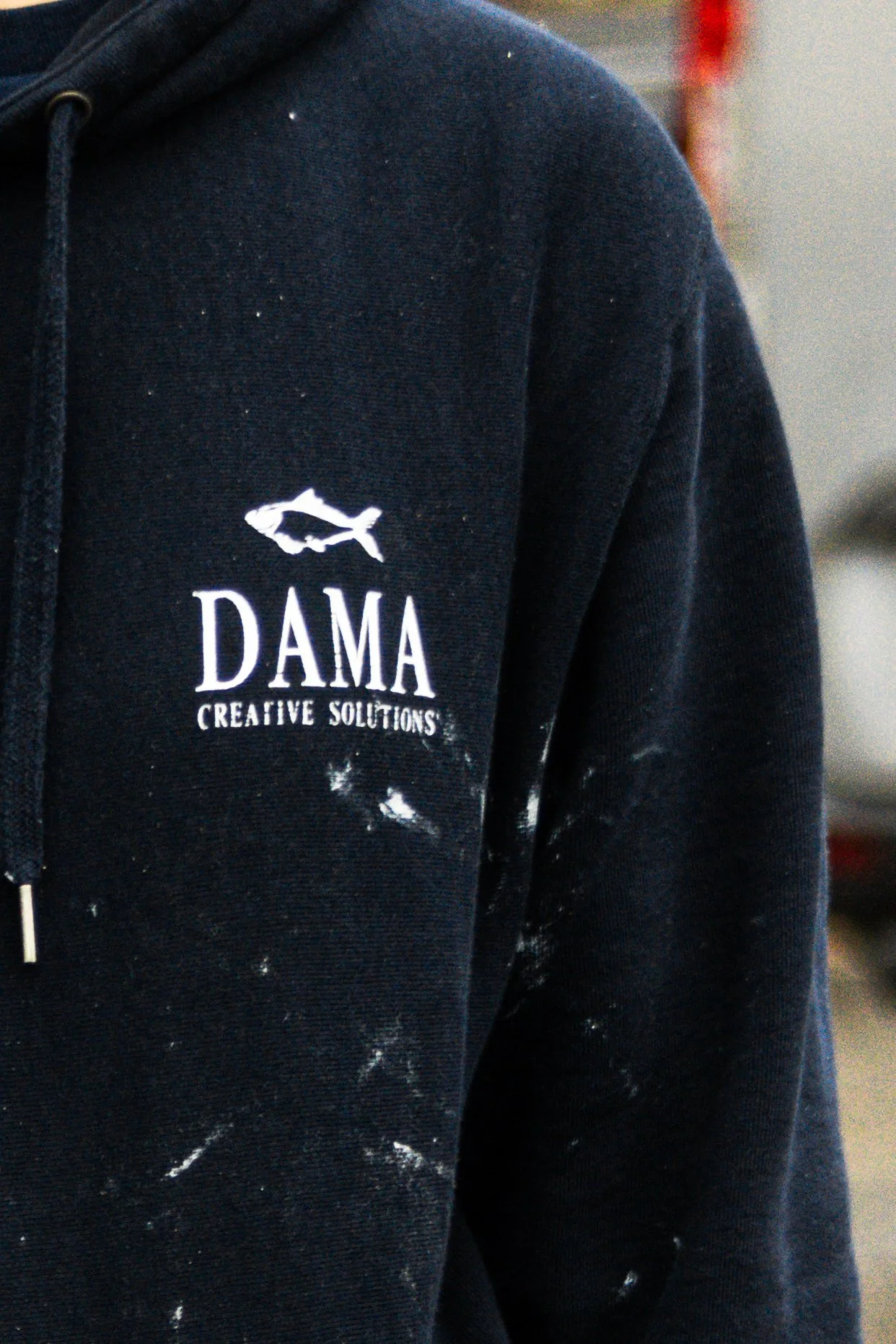 Close-up of a person's black hoodie with a small fish logo and the text 'DAMA CREATIVE SOLUTIONS' on the left chest area, with white paint splatters on the fabric.
