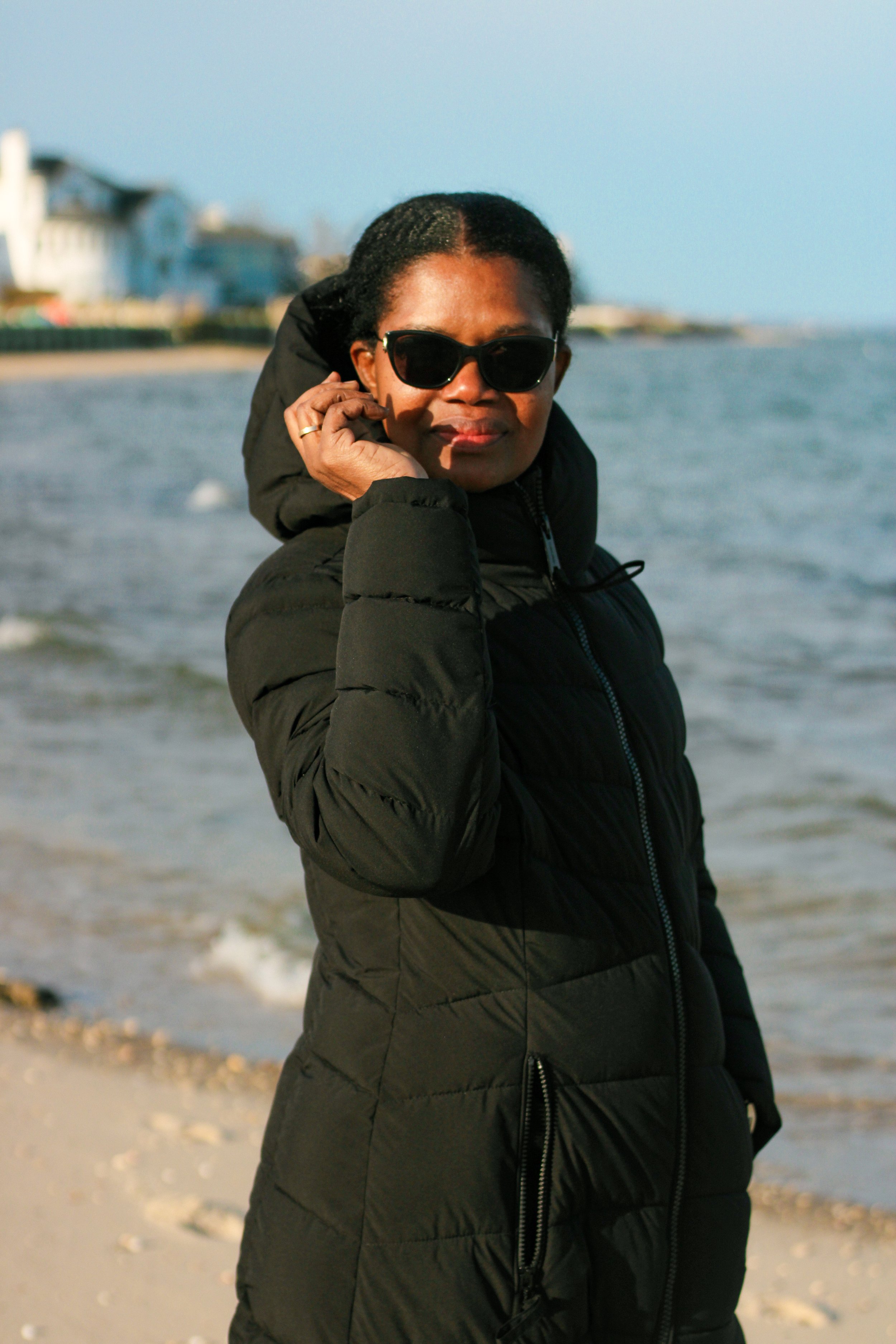 Woman in a black puffer jacket and sunglasses standing at the beach during daytime, with buildings and the ocean in the background.