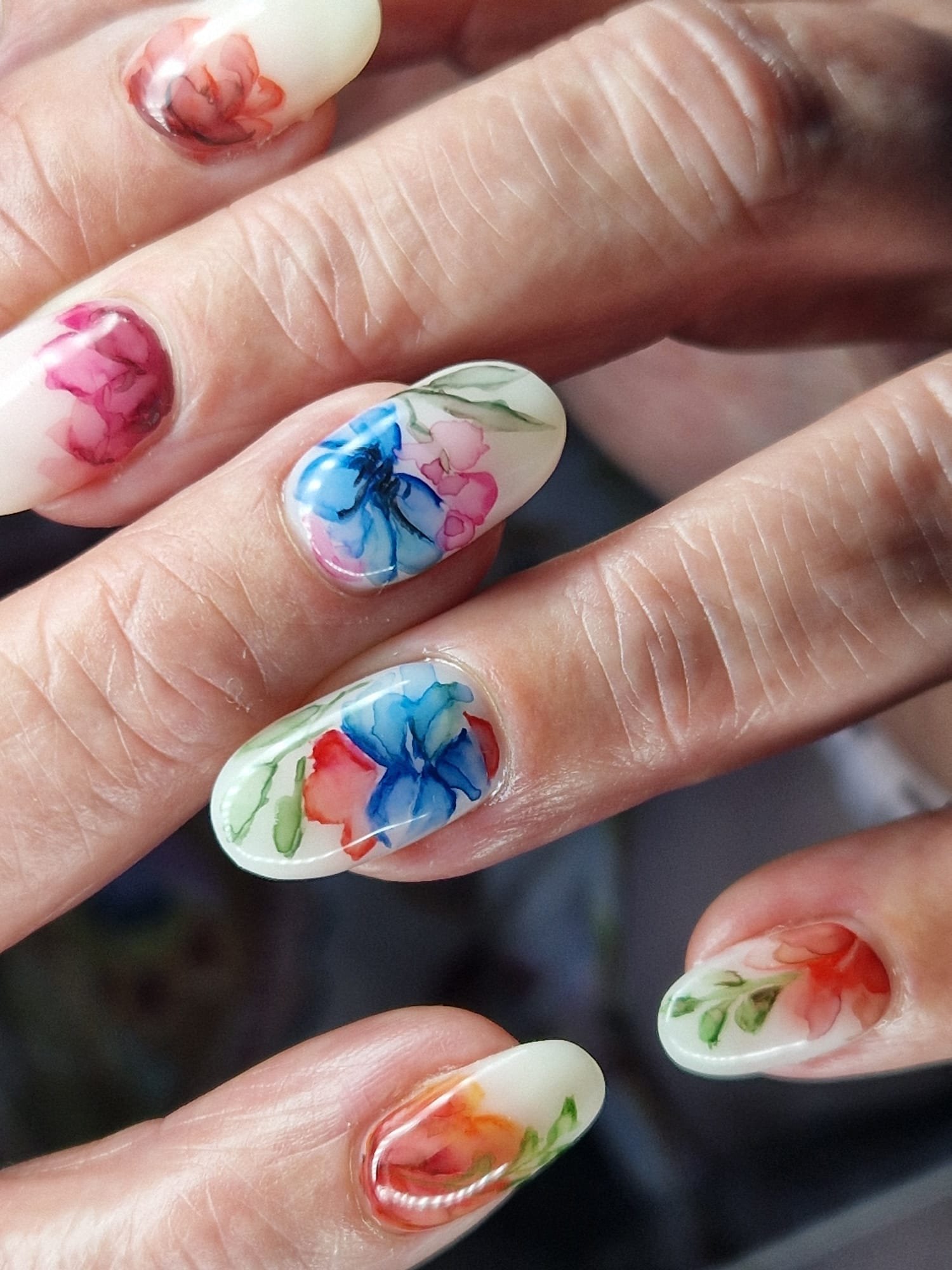 Close-up of several fingers with floral nail art designs featuring colorful flowers in pink, red, blue, and green on a white background.