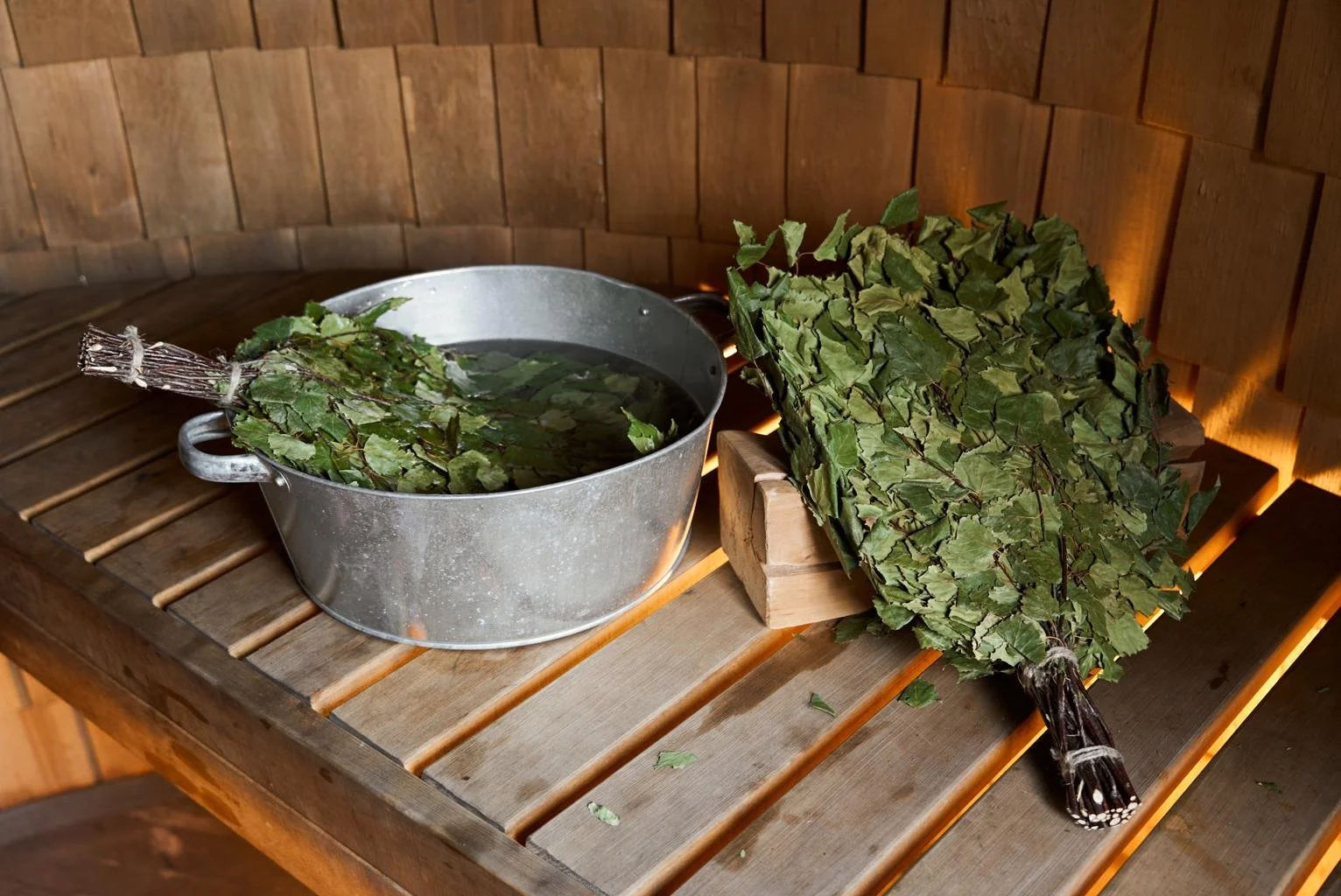 A metal bucket filled with water and green leaves, placed on a wooden surface inside a sauna, with a bundle of dried green leaves resting beside it.
