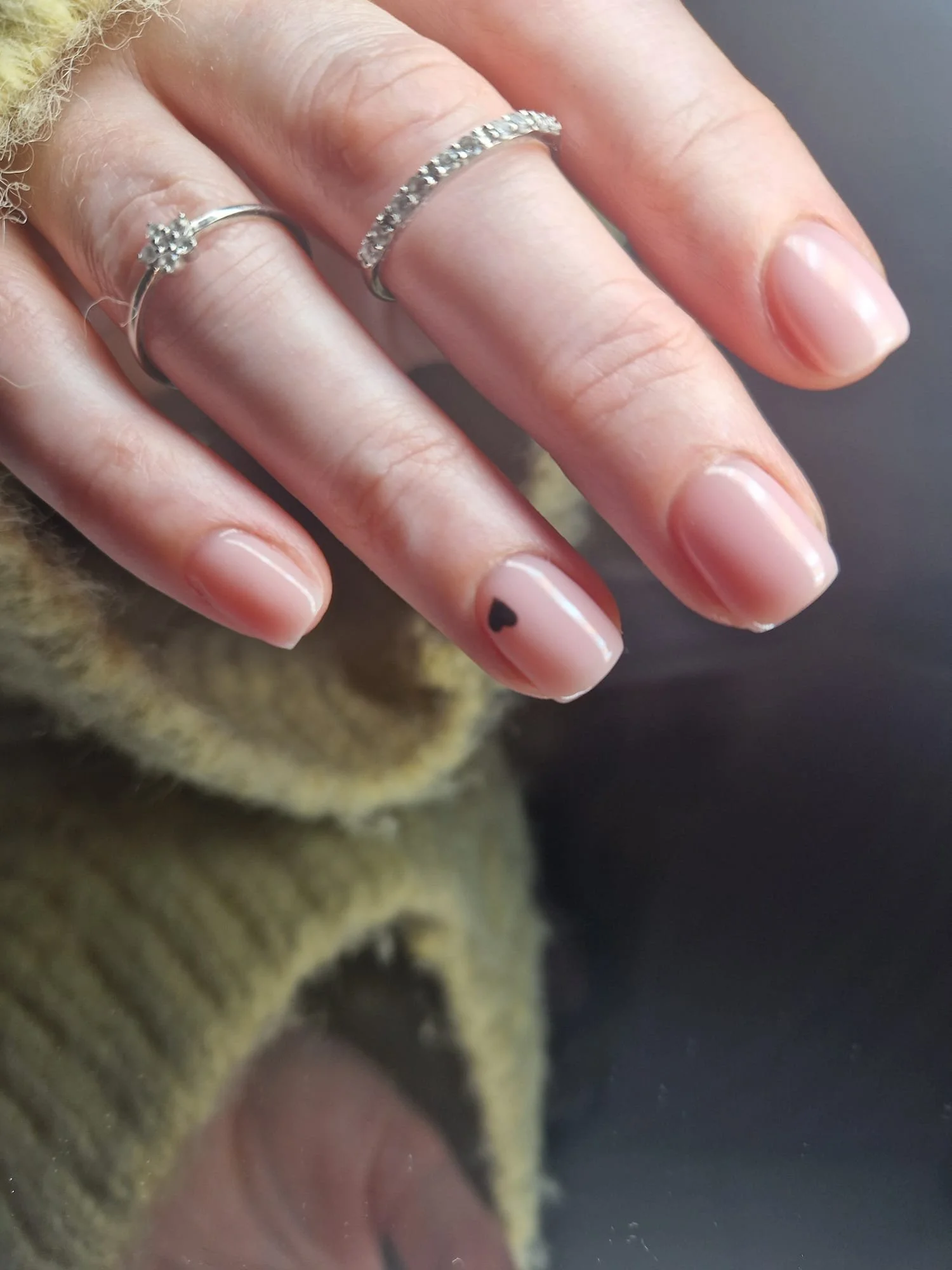 Close-up of a hand with two rings and a small black heart tattoo on the ring finger nail, wearing a mustard-colored knitted sleeve.
