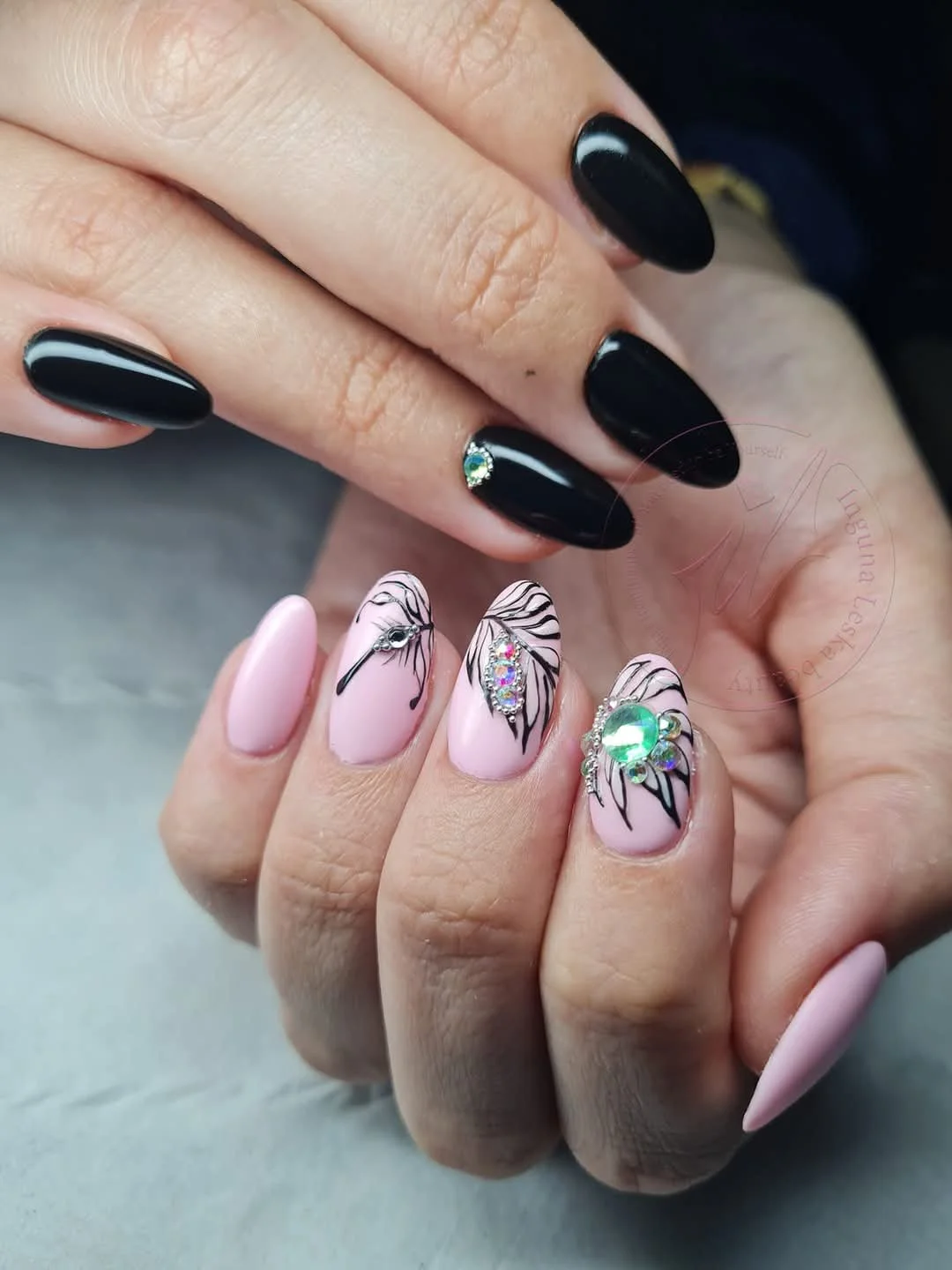 Close-up of hands with manicured nails, featuring black nails with gemstone accents on the index finger and decorated nails with pink polish, black line art, and rhinestone embellishments.