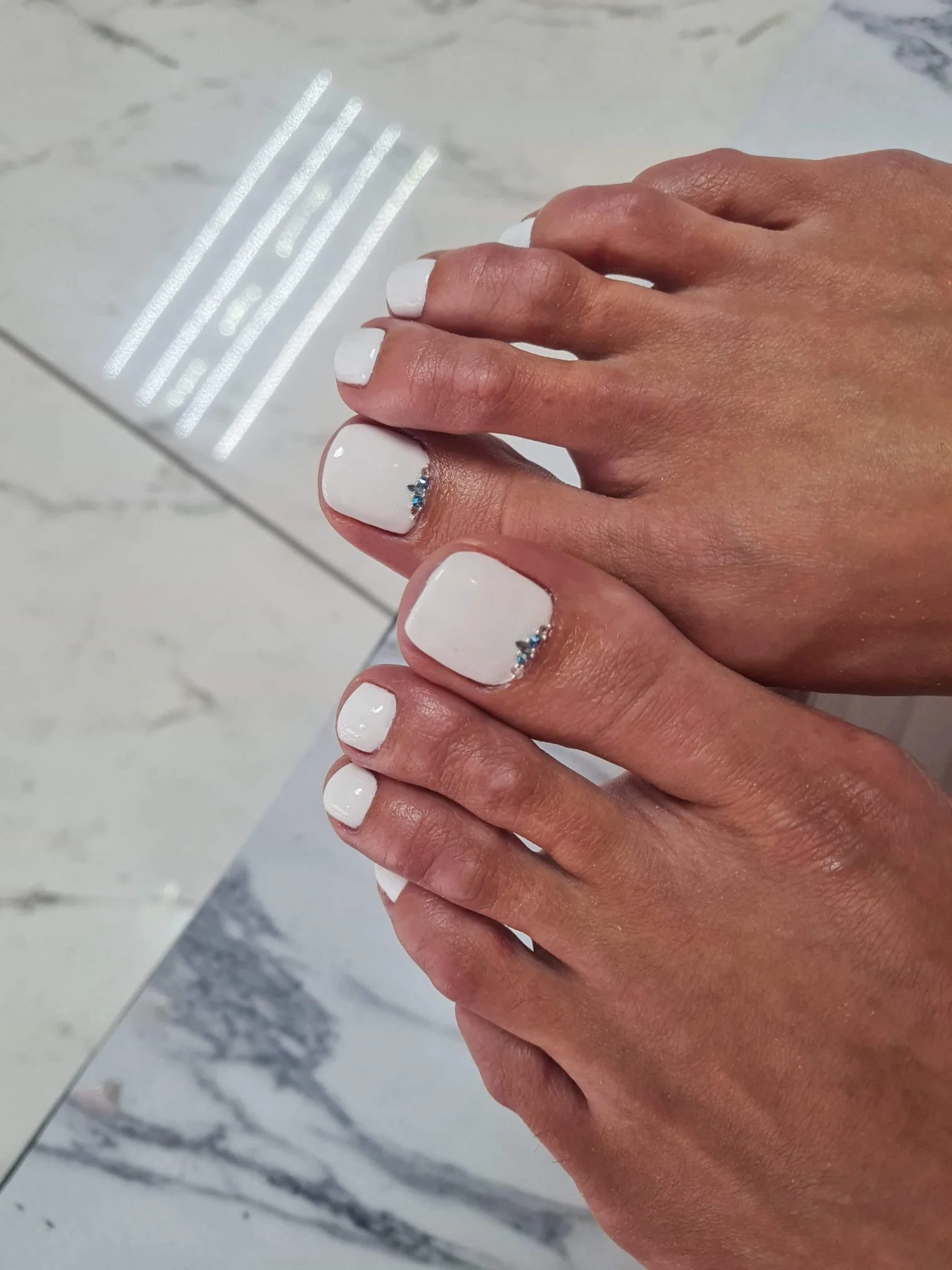 Close-up of a hand with nails painted white and decorated with small blue rhinestones, resting on a marble surface.