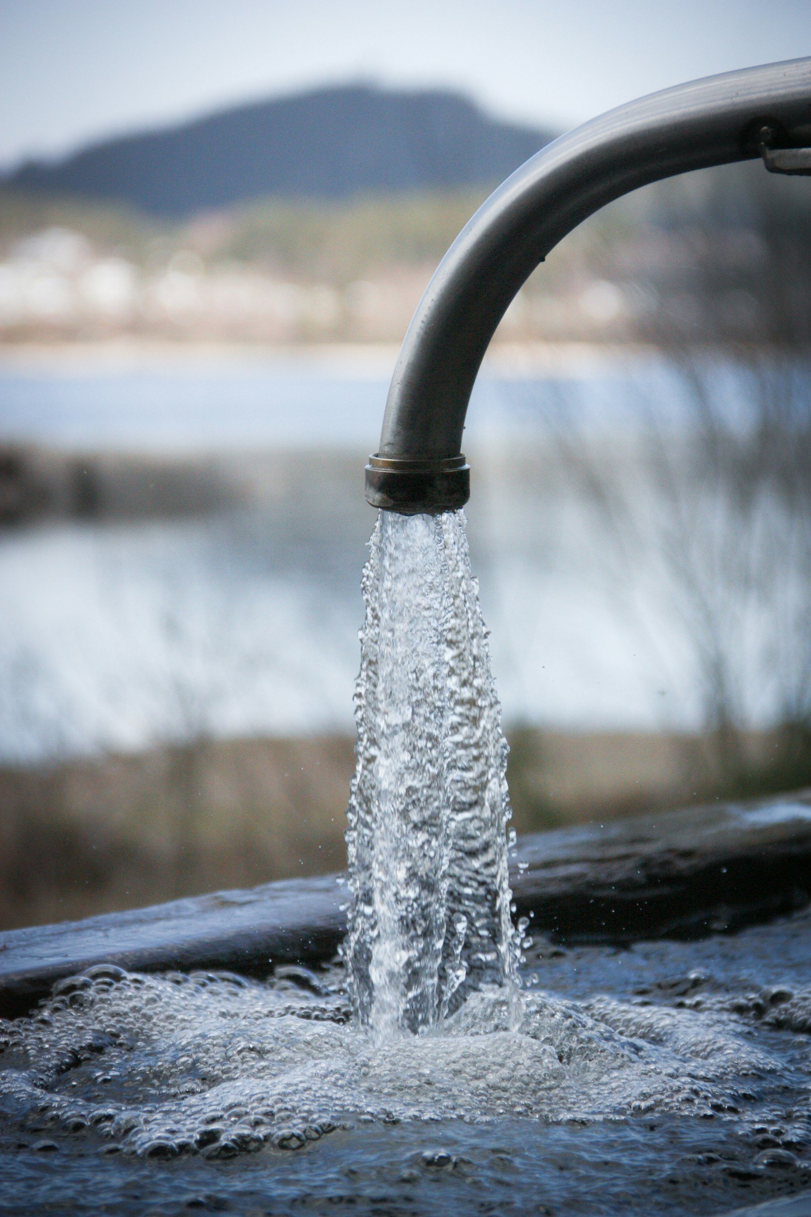 Close-up of a metal outdoor water tap with water flowing from it, with a blurred landscape background including a mountain and trees.
