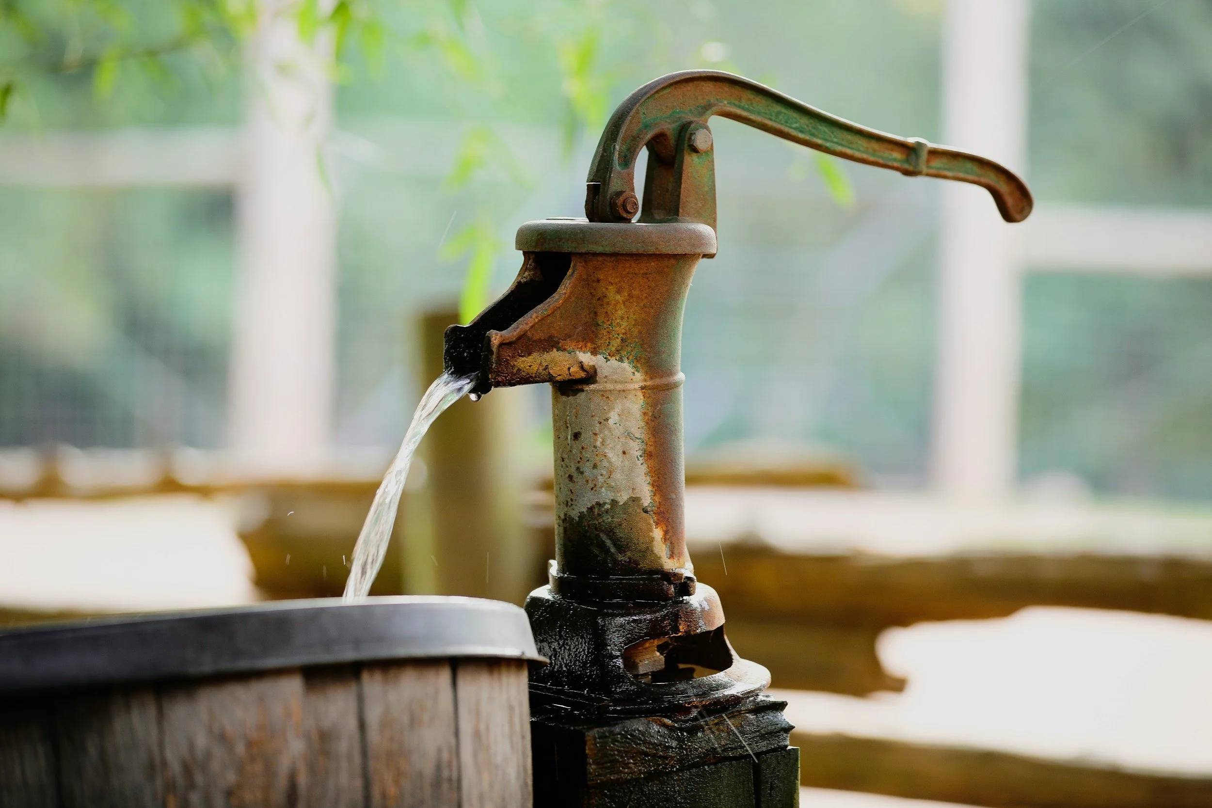 Old rusty hand water pump with water flowing into a wooden basin, outdoors with green blurred background.